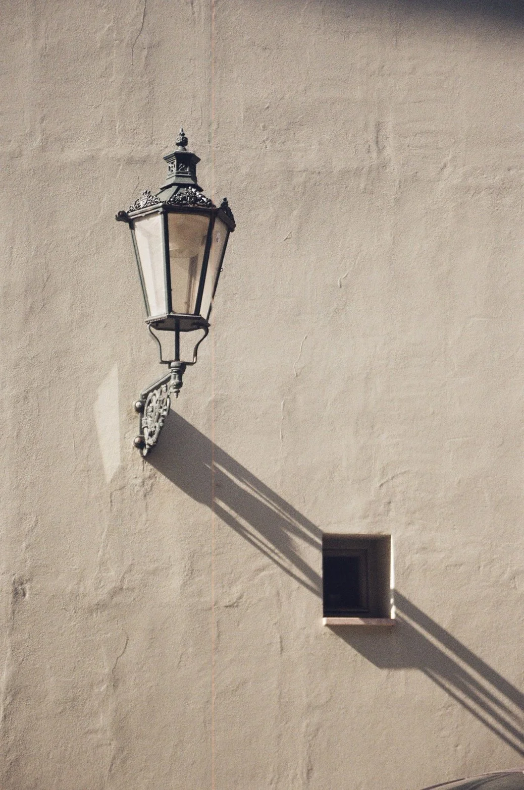 An ornate black metal street lamp casting a shadow on a beige textured wall, with a small square window beneath the shadow.