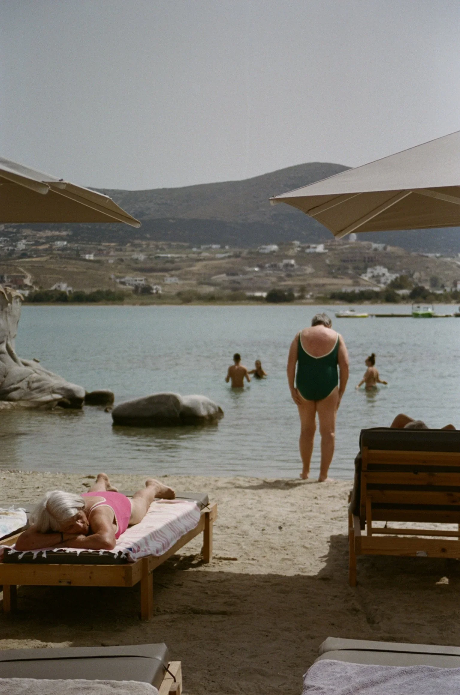 People relaxing on a beach with umbrellas, some swimming in the water, in a scenic coastal area with hills and boats in the background.