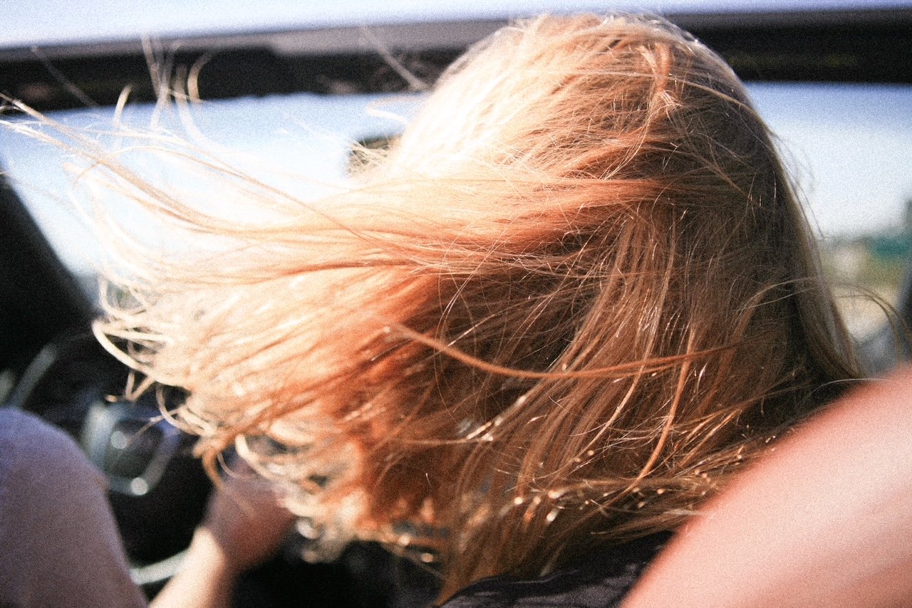 Close-up view of a person's head with long, light brown or blonde hair blowing in the wind, partially obscuring their face, inside a moving vehicle.