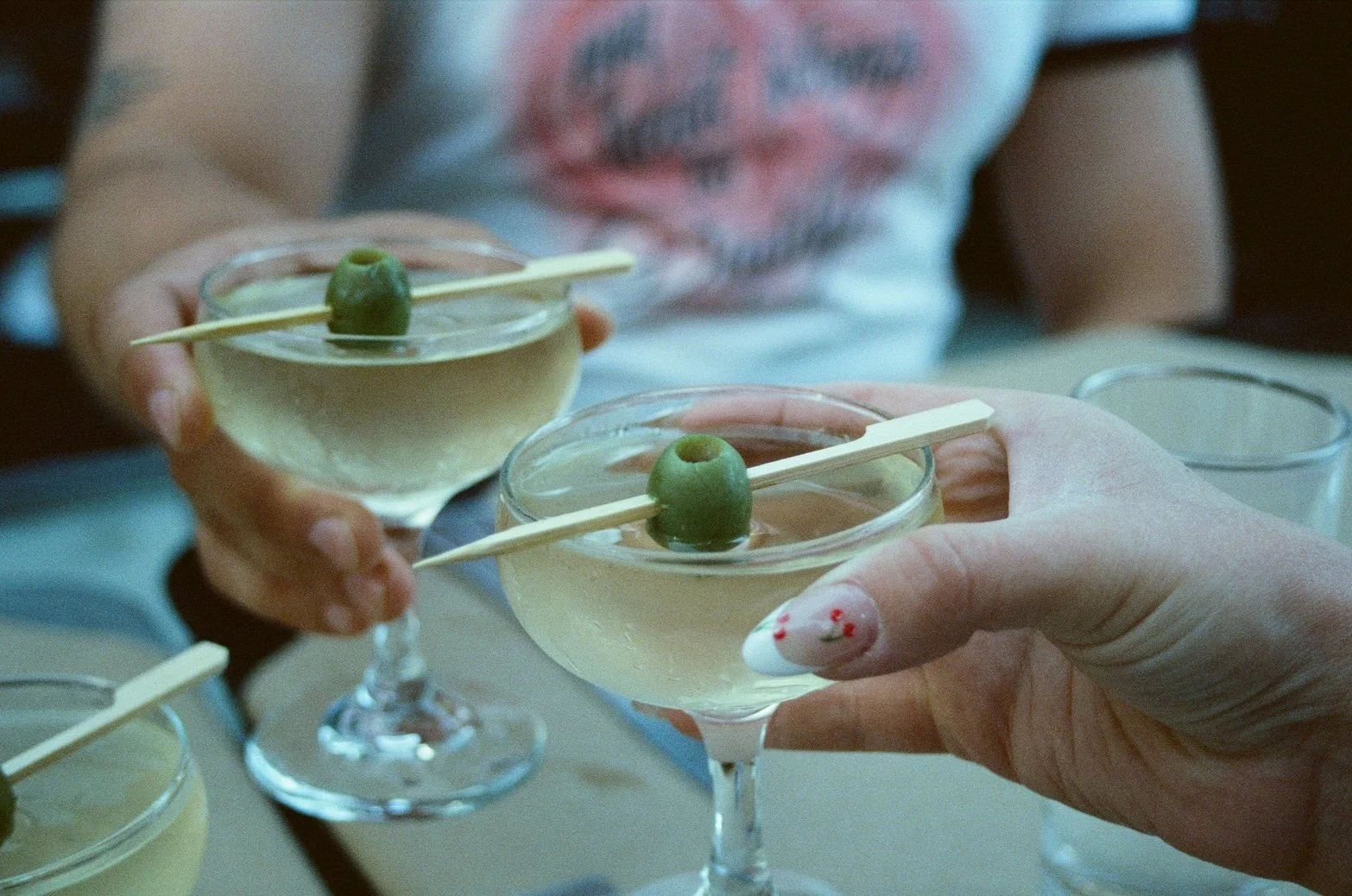 Two people holding cocktails garnished with green olives, with one person's fingernail decorated with a floral design, in a social setting.