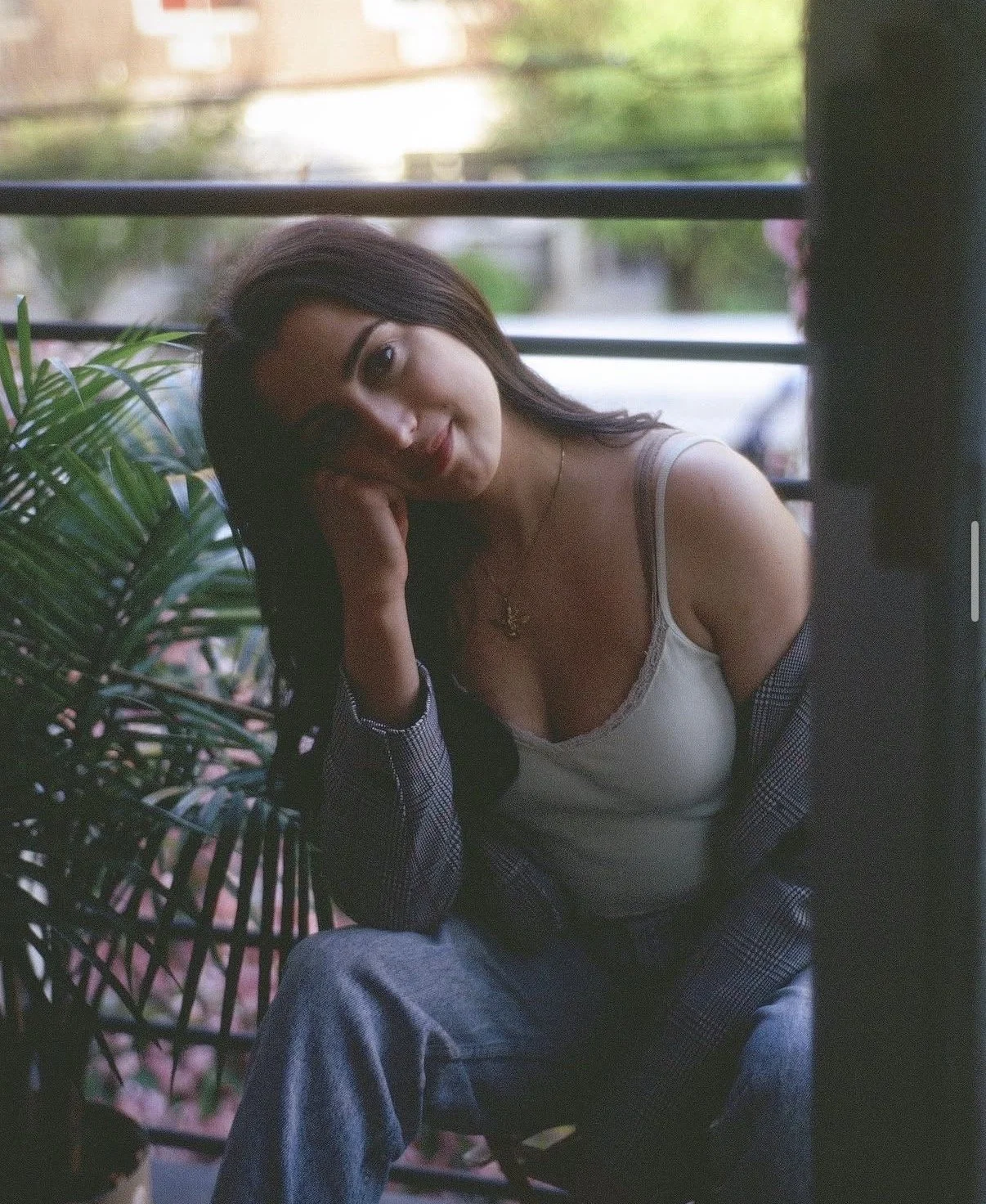 A young woman sitting outdoors near a window, with green plants and trees in the background. She has long dark hair, is smiling, and resting her head on her hand. She is wearing a white tank top, a checkered shirt, and jeans.