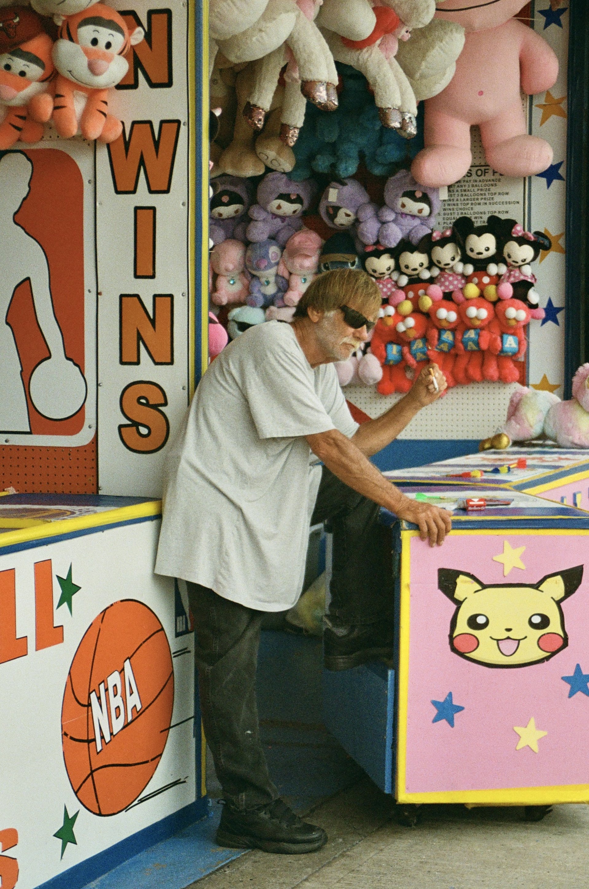 A man wearing sunglasses and a white t-shirt sitting at a carnival game booth with plush toys and stuffed animals displayed behind. The booth features colorful signs with cartoon characters and sports-themed decorations.