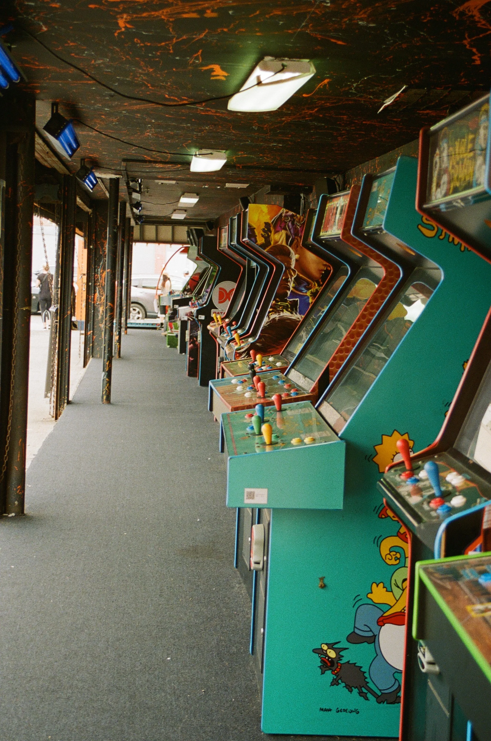 Several vintage arcade game machines with colorful artwork featuring characters from The Simpsons lined up inside a bar or arcade with black marble walls and a dark ceiling.