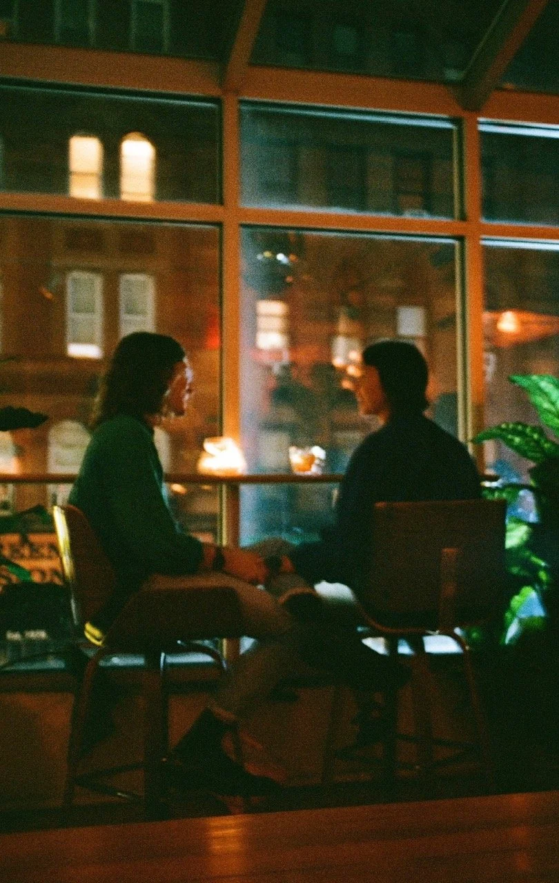 Two people sitting and talking at a table in a dimly lit restaurant or cafe, viewed through large glass windows with a reflection of the interior. They are holding hands or touching.