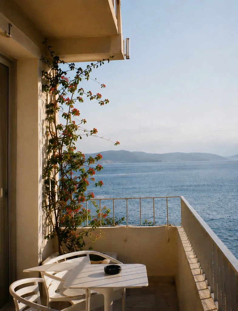 Balcony overlooking a body of water with distant landforms, white outdoor furniture including a table, two chairs, a bowl, and a climbing plant with red flowers on the left side.