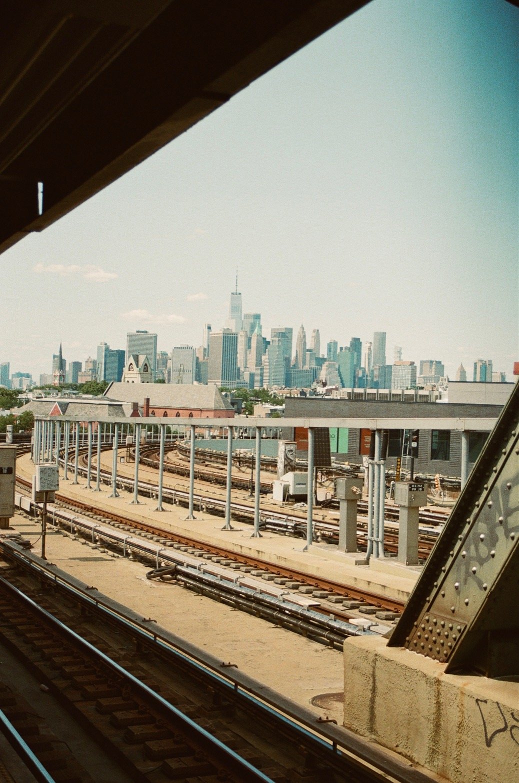 View of the Manhattan skyline from a train track platform, including tracks, infrastructure, and the One World Trade Center in the distance.