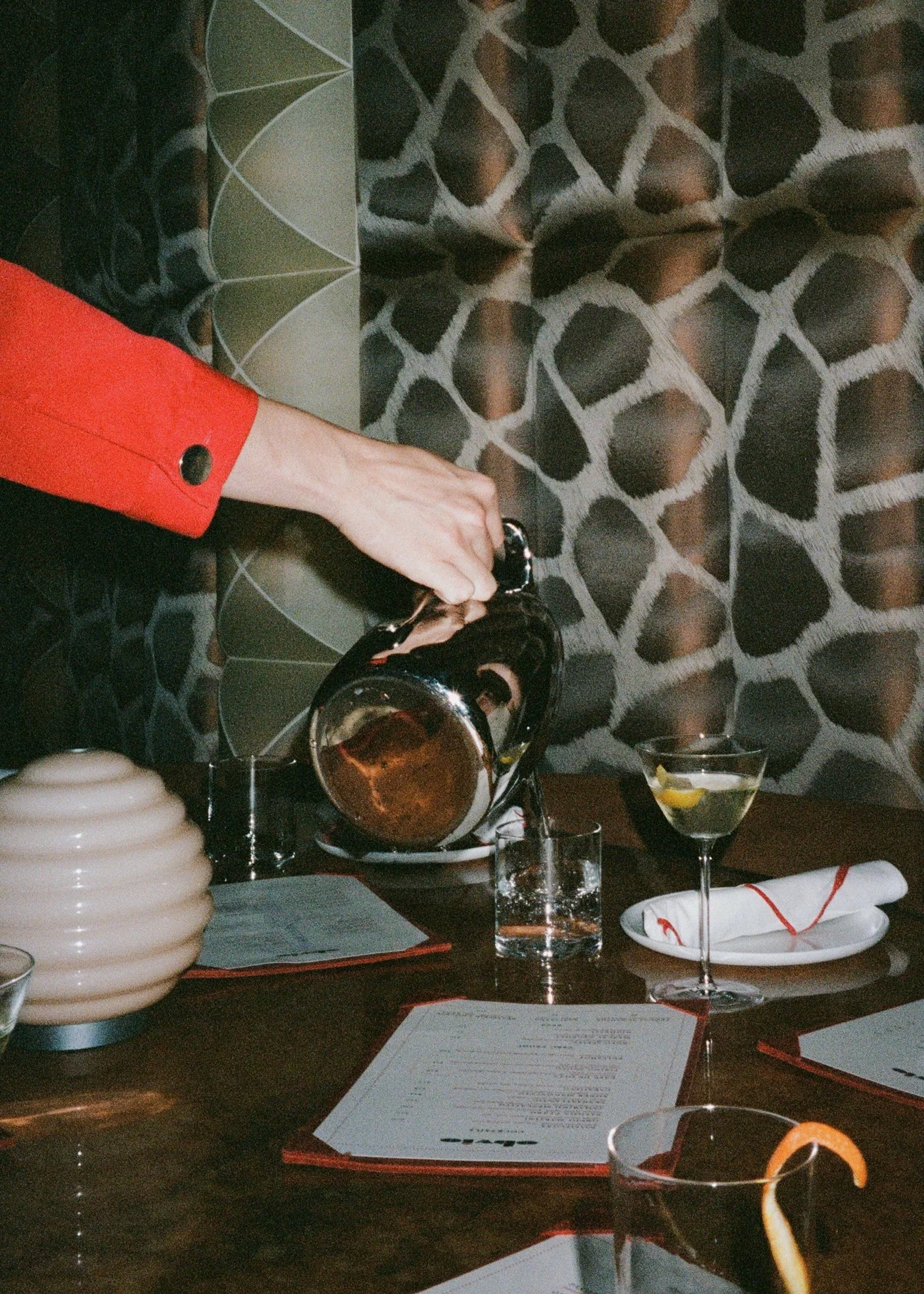 A person wearing a red sleeve pours a drink from a teapot into a glass on a dark wooden table, with various drinks, menus, glasses, a candle, and a decorative object on the table, against a patterned black and white background.
