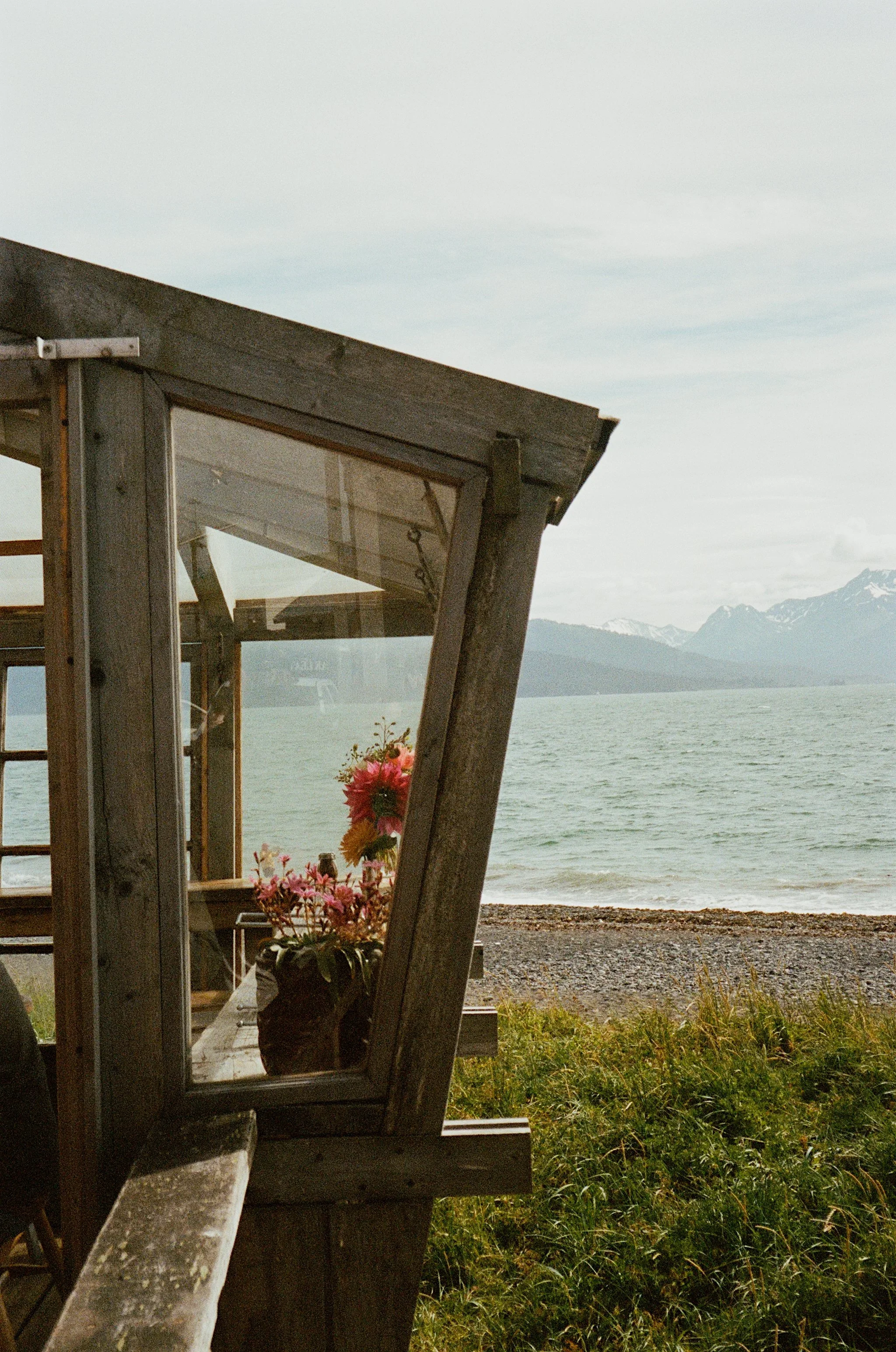 A view of a lakeside with a wooden structure, flowers in a pot, and mountains in the background under a cloudy sky.