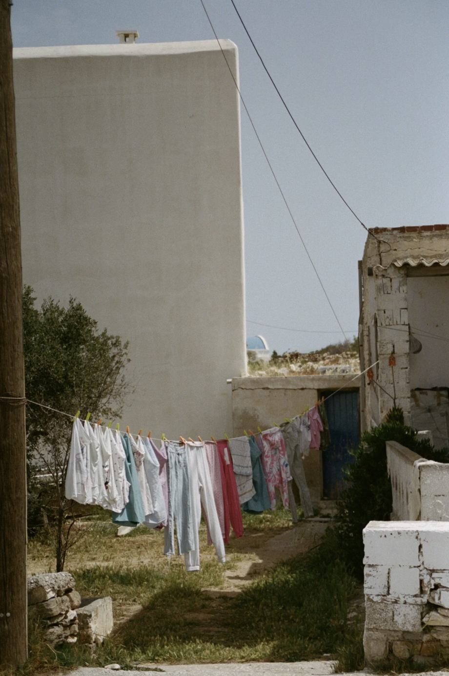 Clothes hanging on a clothesline outside between wooden posts and buildings, with some bushes and a dirt path below, in a sunny outdoor scene.
