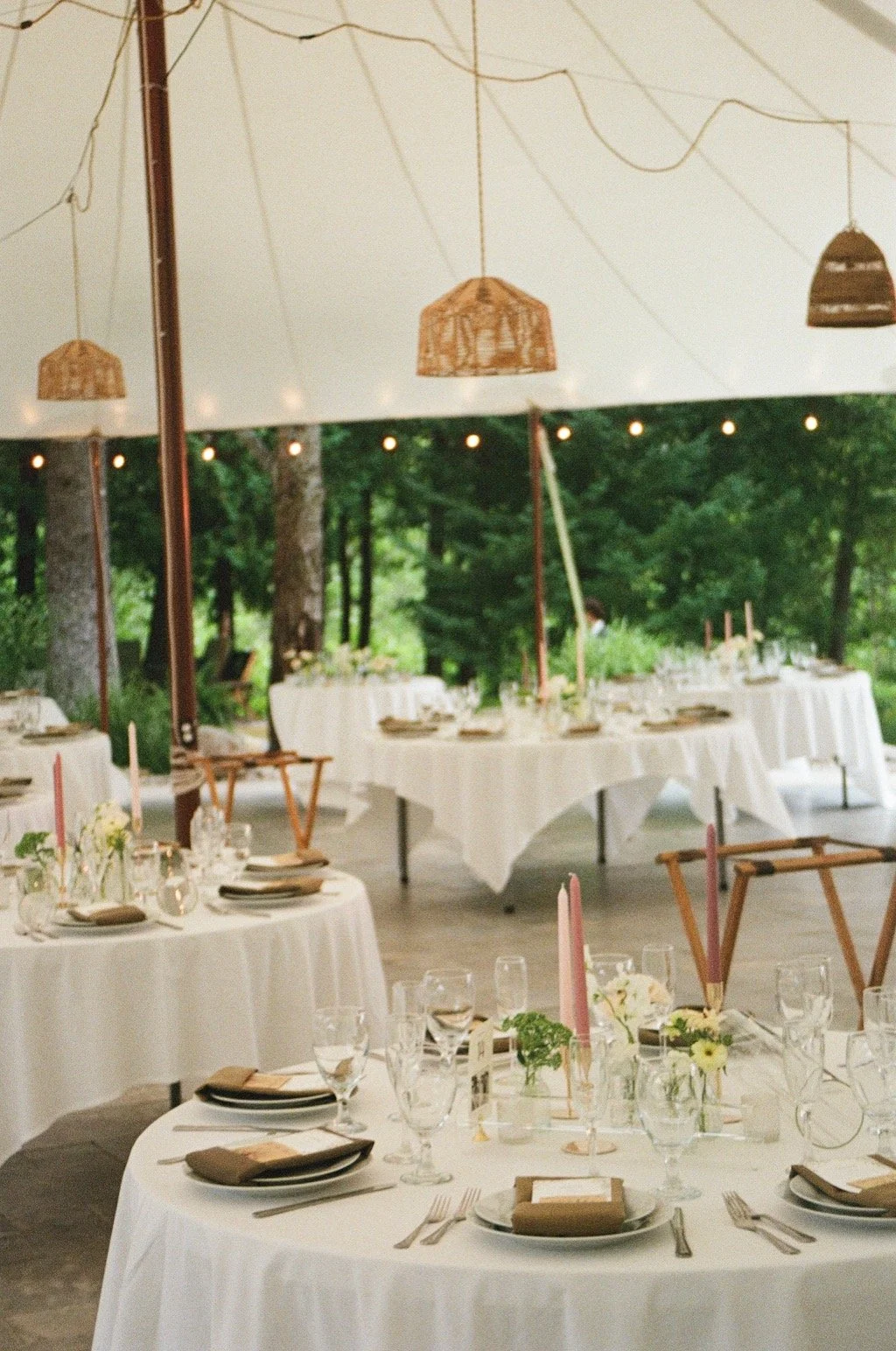 Decorated outdoor event space with round tables covered in white tablecloths, set with plates, glasses, napkins, and floral centerpieces, under a tent with hanging wicker lamps and string lights in a green, wooded area. wedding, bridal, place setting