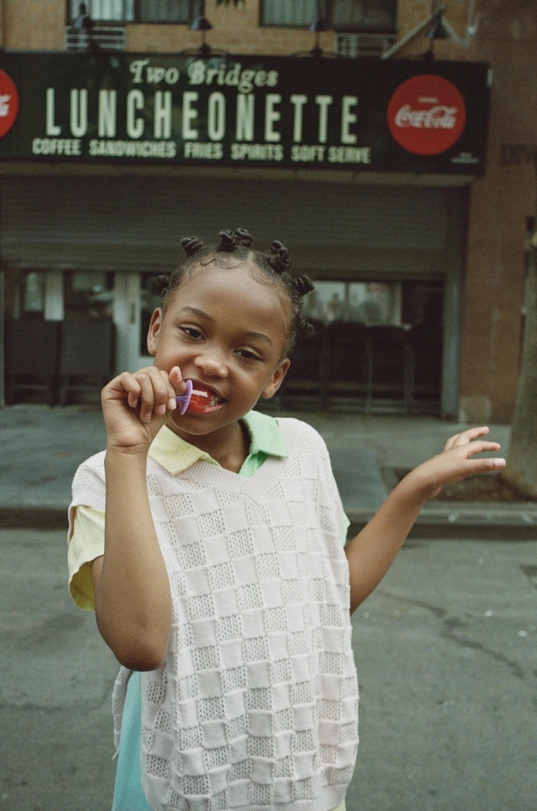 A young girl with Bantu knots hairstyle smiling and holding a pacifier in front of a storefront with a sign that reads "Two Bridges Luncheonette," advertising coffee, sandwiches, fries, sports, and soft serve.