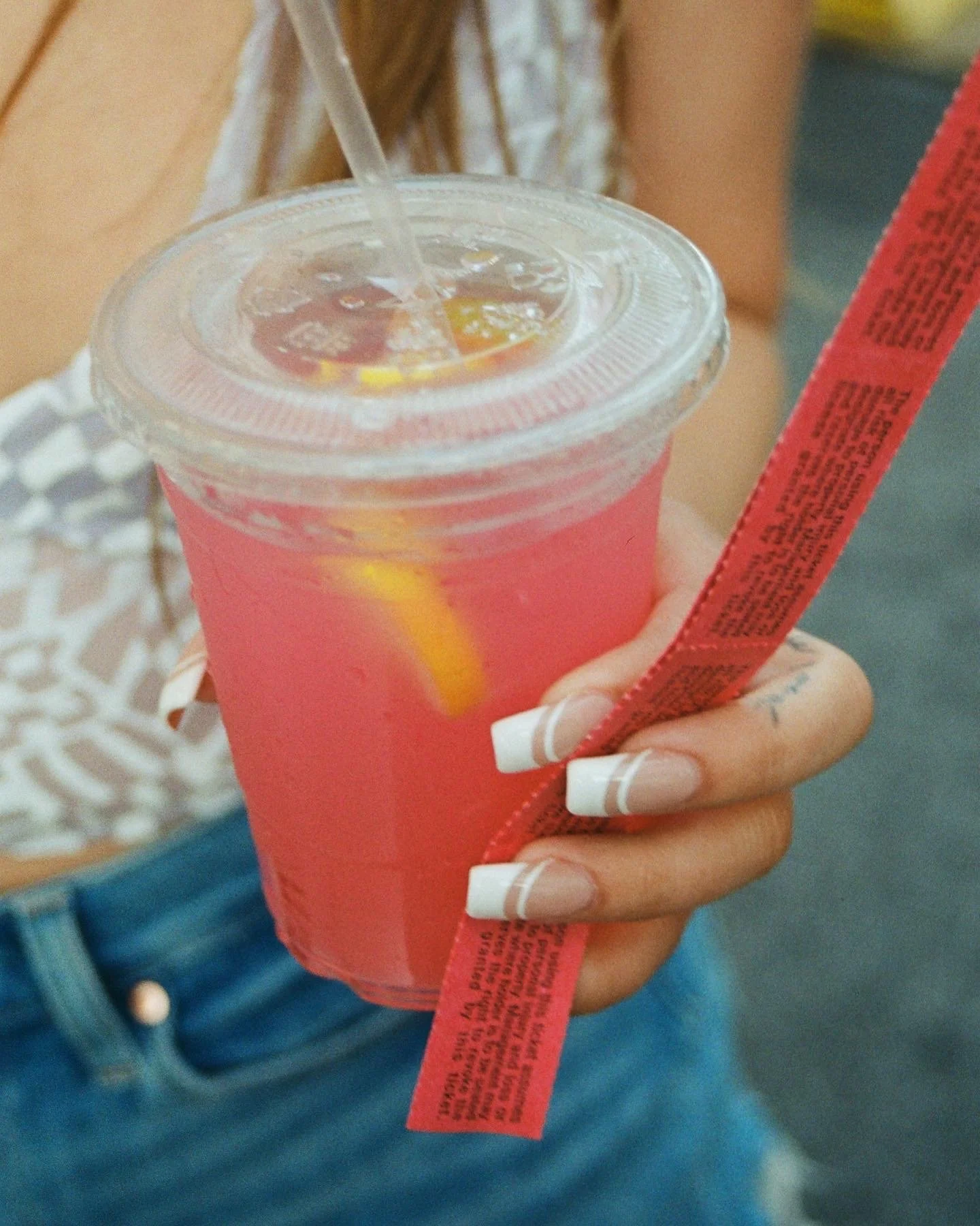Close-up of a person holding a pink cold beverage with a yellow straw in a clear plastic cup with a sealed lid. The person has neatly manicured white French tips and is wearing a blue denim jacket and a ring on their hand.