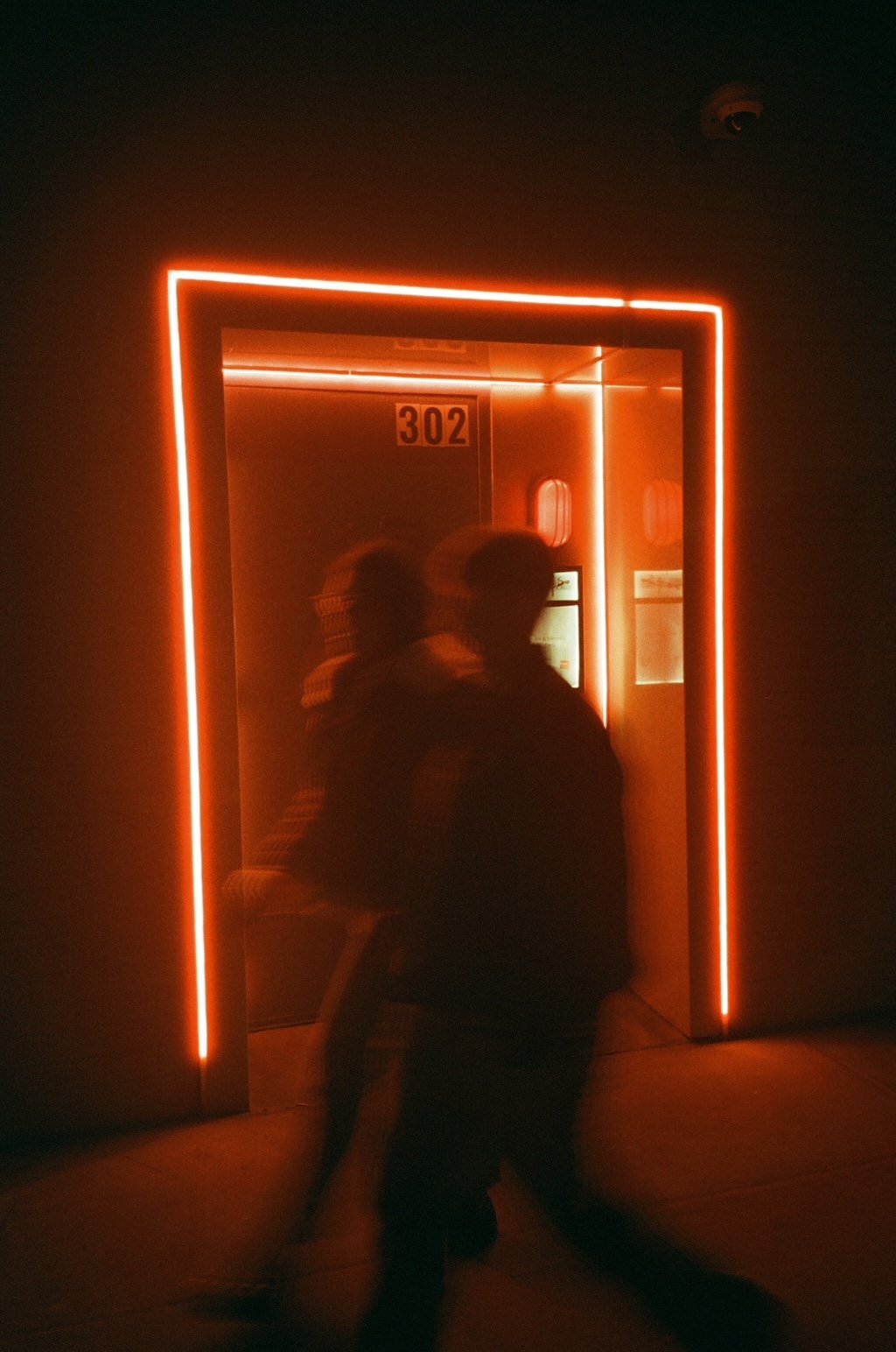 Two people walking past a doorway with red neon lights and a sign that reads '302'.
