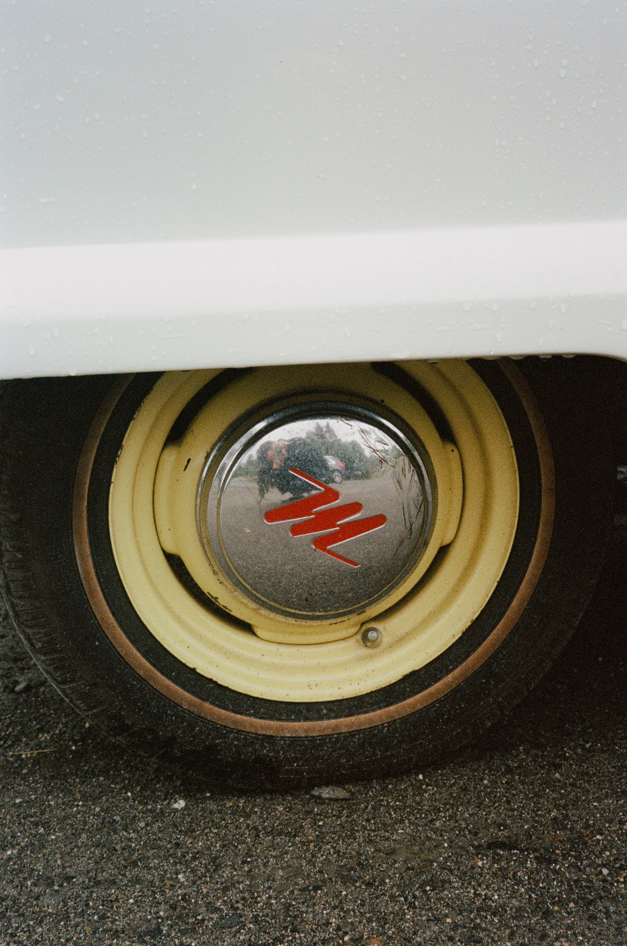 Close-up of a vintage wheel with a cream-colored hubcap and black tire, reflecting a person and a car parked outside on a cloudy day.