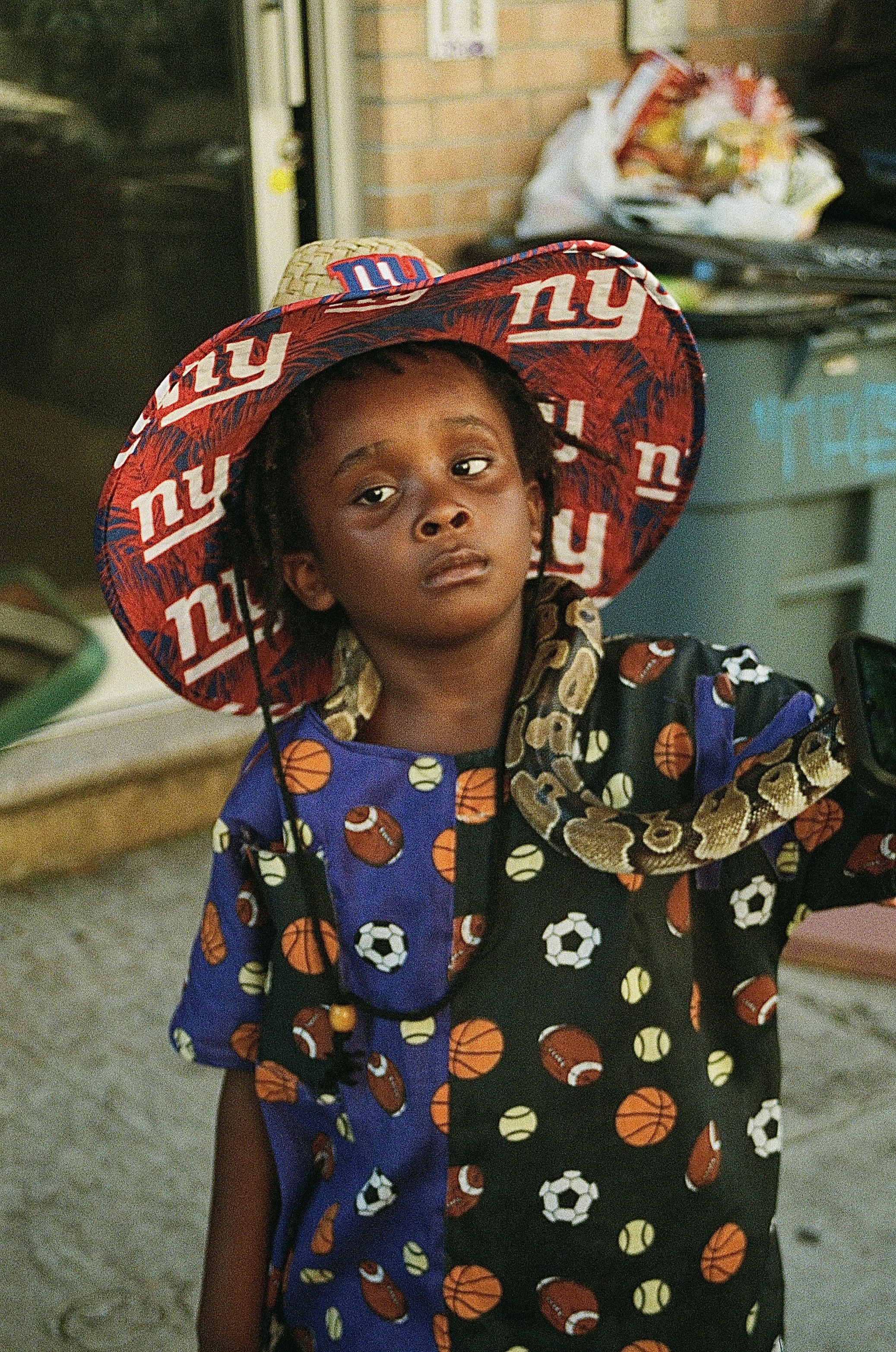 A young child wearing a large New York Giants hat and a football-themed shirt with various sports balls.