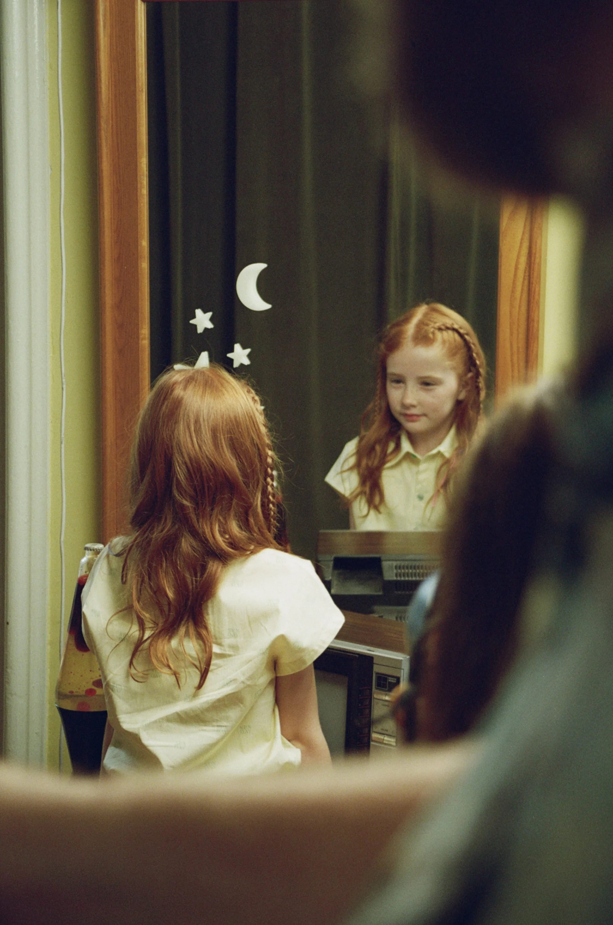 A young girl with red hair and a braid looking at herself in a mirror decorated with moon and stars, while a person with gray hair is in the foreground.