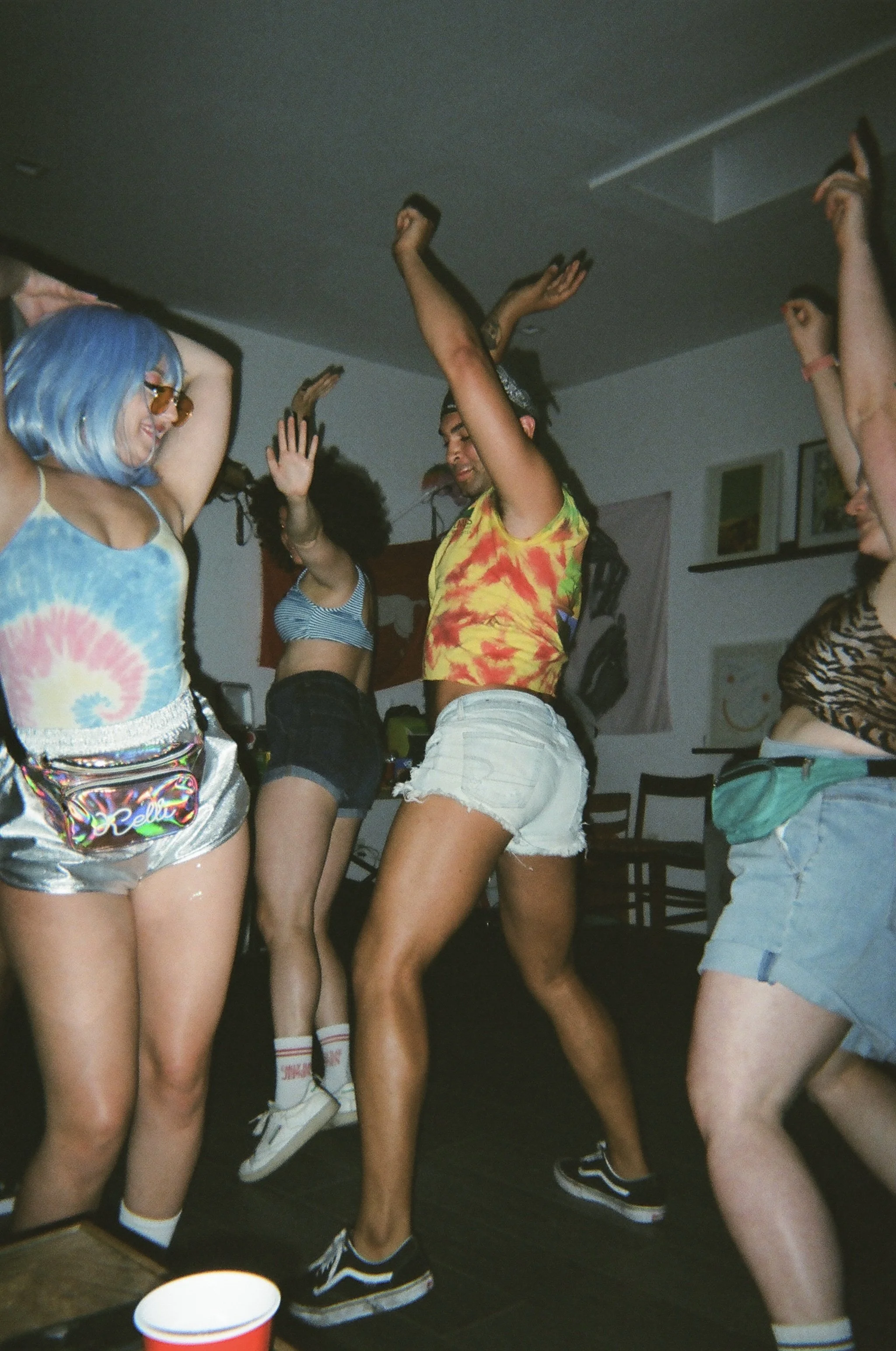 Several people dancing at a party, showing their legs and arms, with a person in a tie-dye top, shorts, and sneakers, and a person with long hair in a tie-dye shirt.