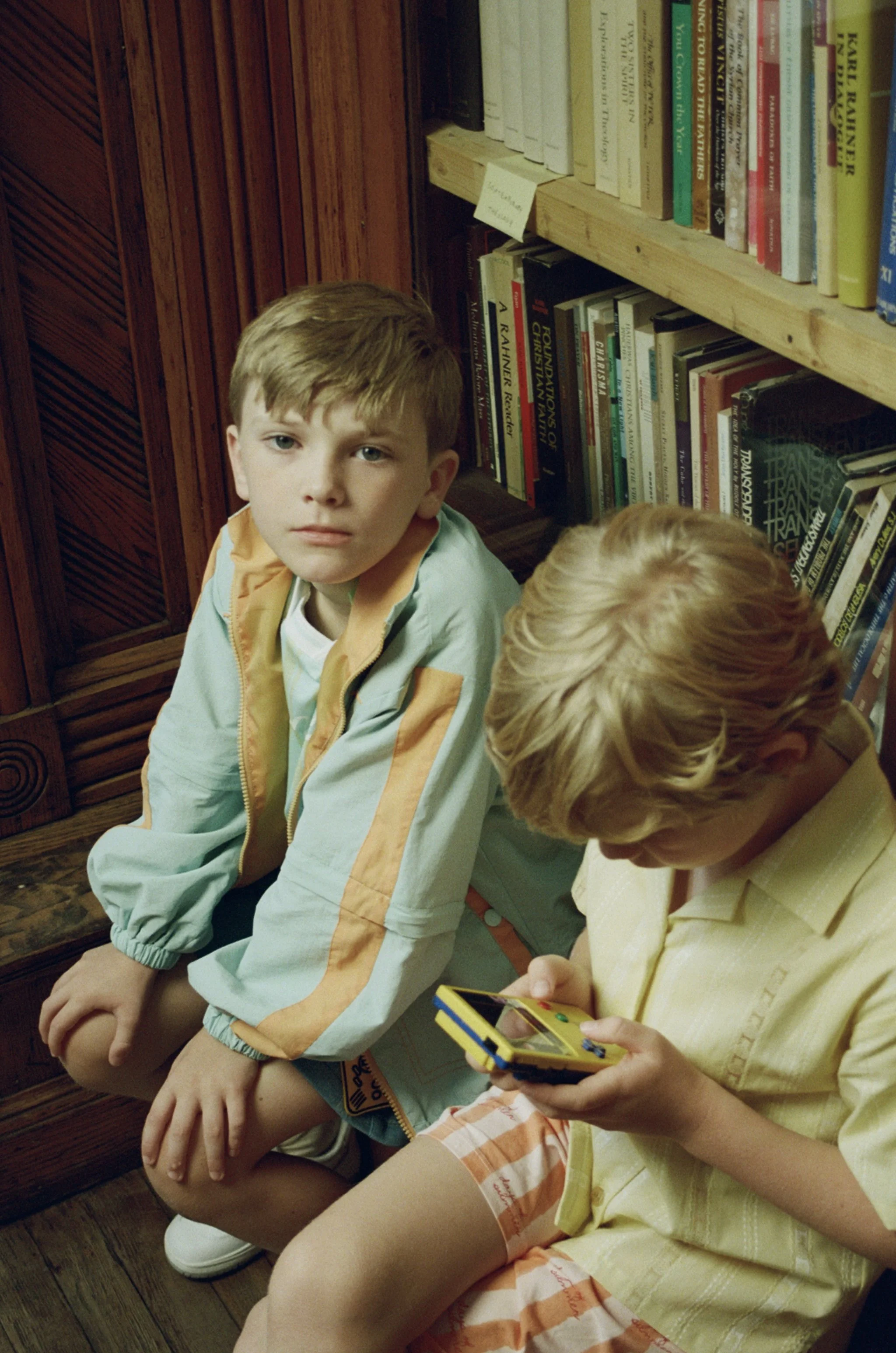 Two young boys sitting on the floor in front of a bookshelf, one looking at the camera with a serious expression, the other holding a yellow handheld electronic game.