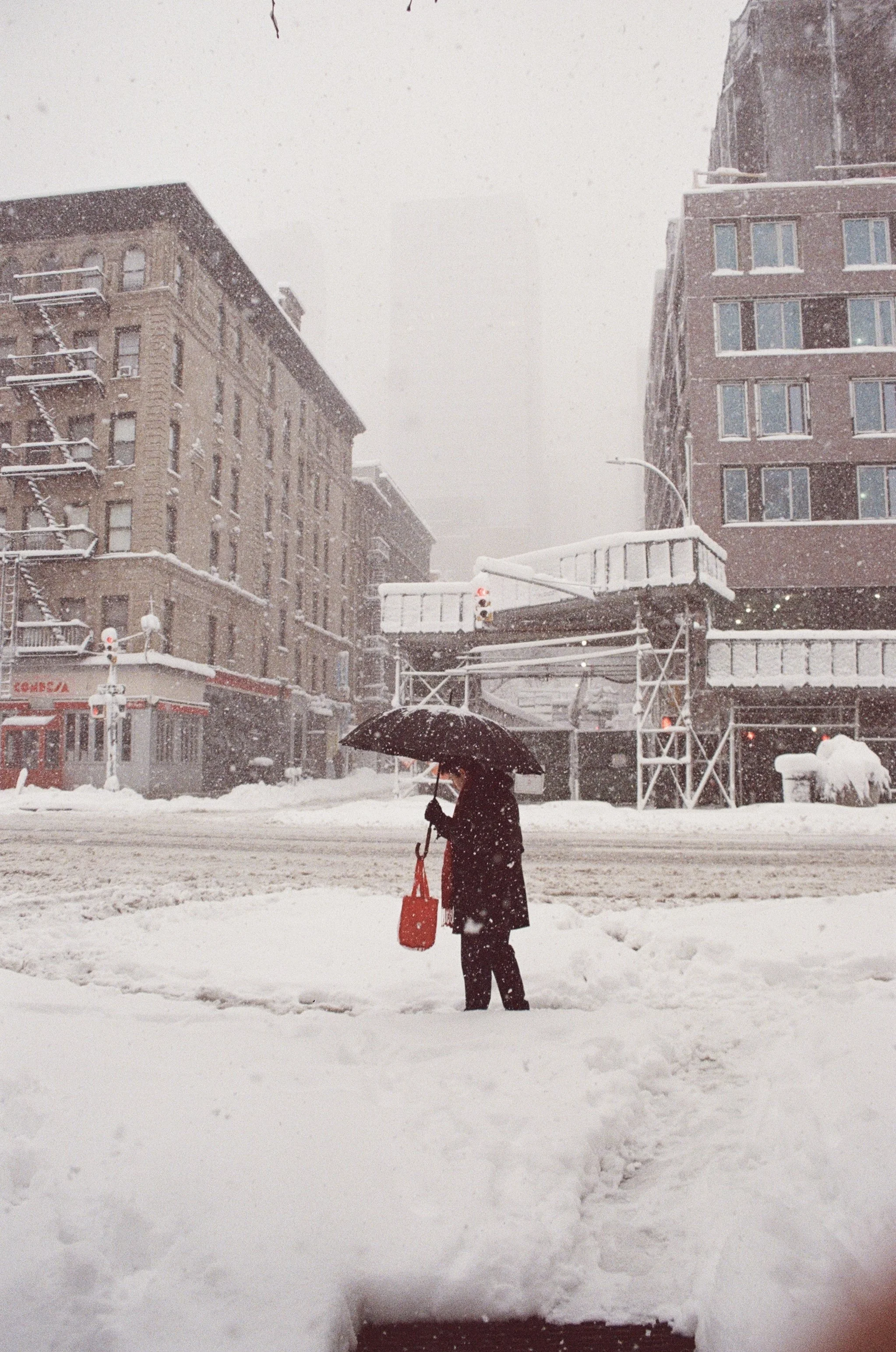 A person standing on a snow-covered street holding a red bag and an umbrella during a snowstorm, with snow falling heavily amidst city buildings.