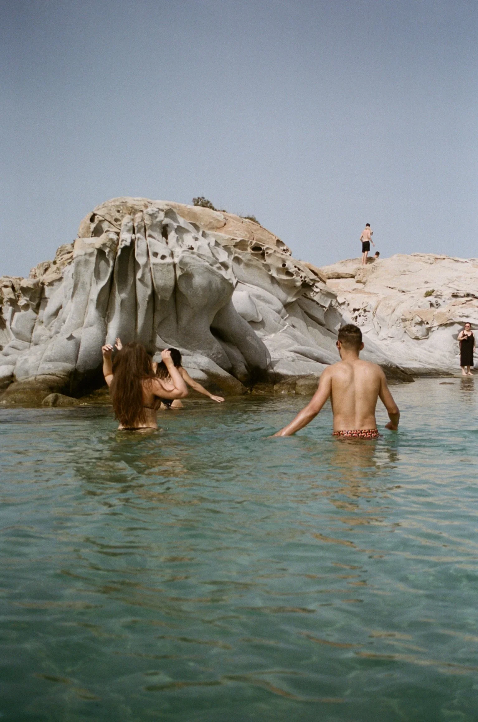 People swimming in clear water near a large, textured rock formation with a person standing on it and others nearby, under a clear sky.