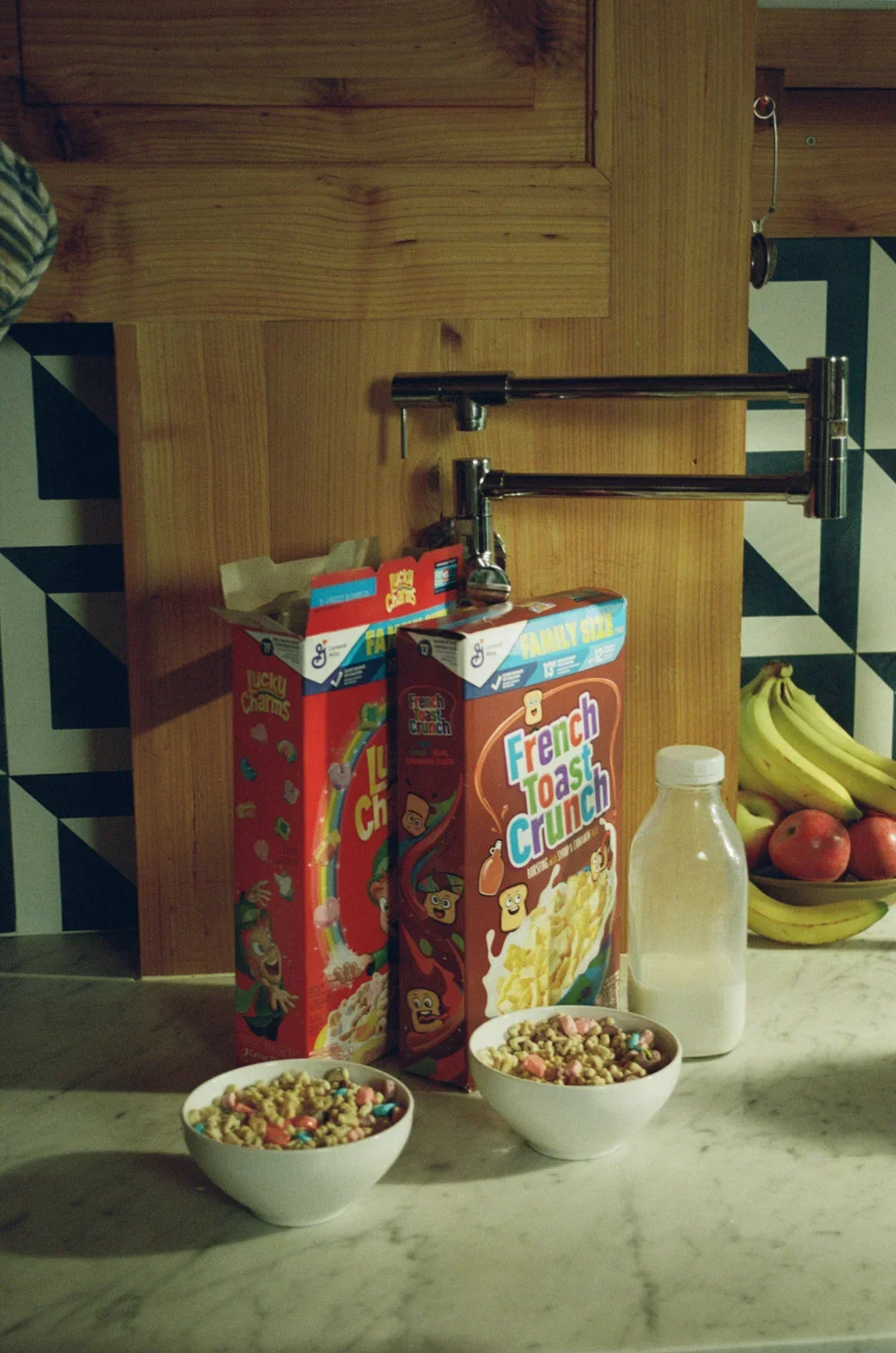 A kitchen countertop with two bowls of cereal, a carton of French Toast Crunch cereal, a milk bottle, a bunch of bananas, and some apples. There is a wooden backsplash and patterned black-and-white tiles in the background.