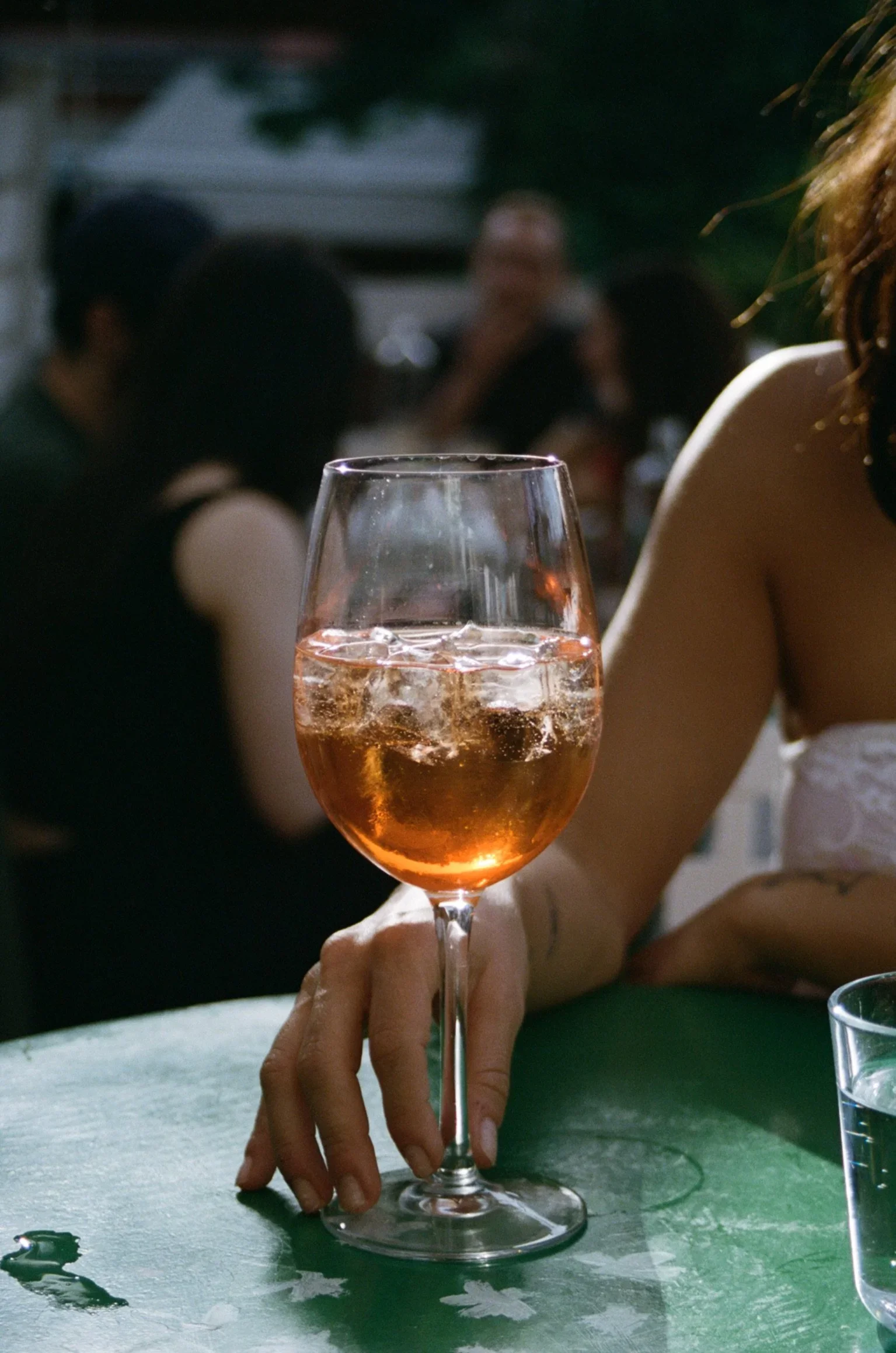 A hand holding a glass of pink-colored drink with ice, with a blurry background showing a woman and other people at a social gathering.
