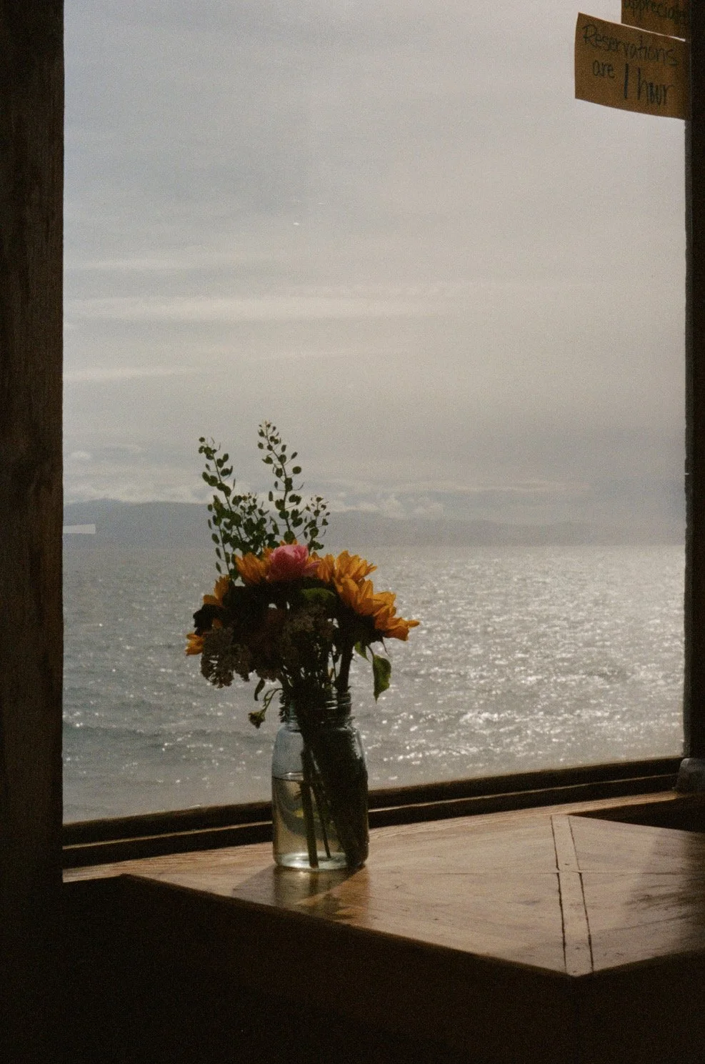 A glass jar with a bouquet of yellow, pink, and purple flowers, along with green leaves and branches, sits on a wooden windowsill overlooking a large body of water under a cloudy sky.