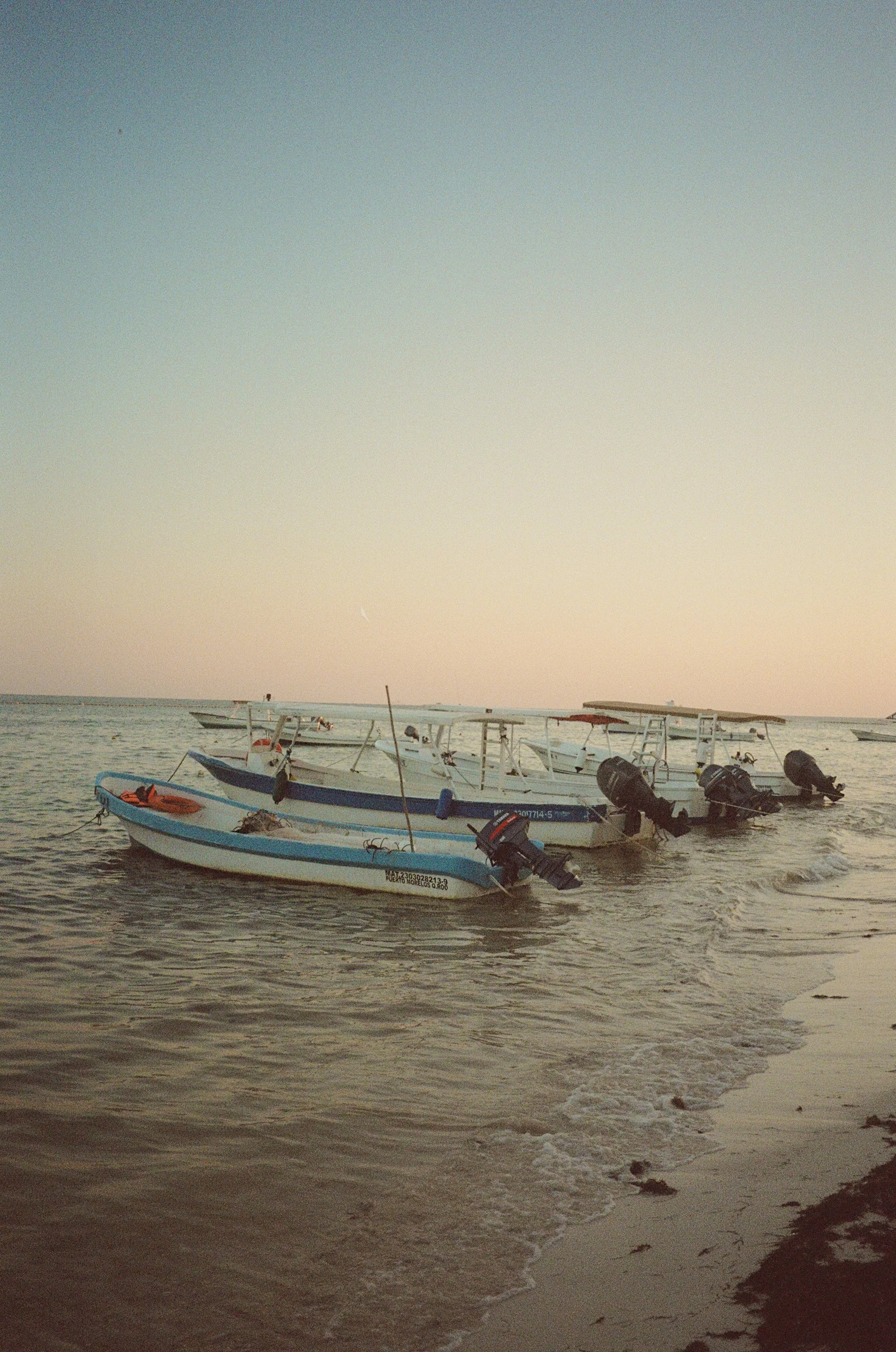 Several small boats anchored near the shore at sunset or sunrise, with gentle waves lapping against the sand and a pastel sky.
