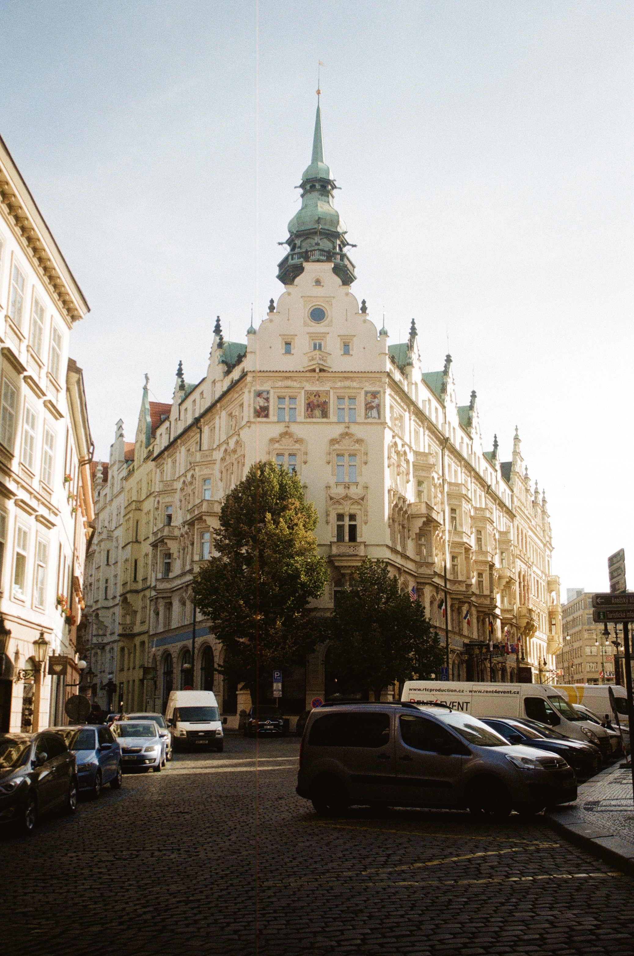 A historic building with ornate architecture and a tall spire, located at the intersection of city streets with parked cars and trees in the foreground, under a clear sky.
