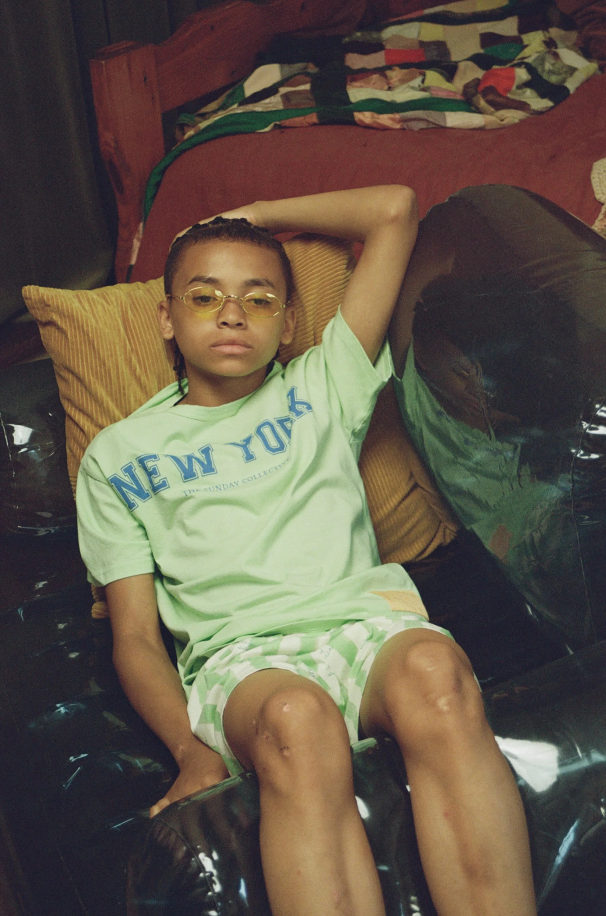 A young boy with short hair, glasses, wearing a light green shirt with blue letters and matching shorts, laying on a yellow pillow on a black couch, with his eyes closed and arm behind his head.
