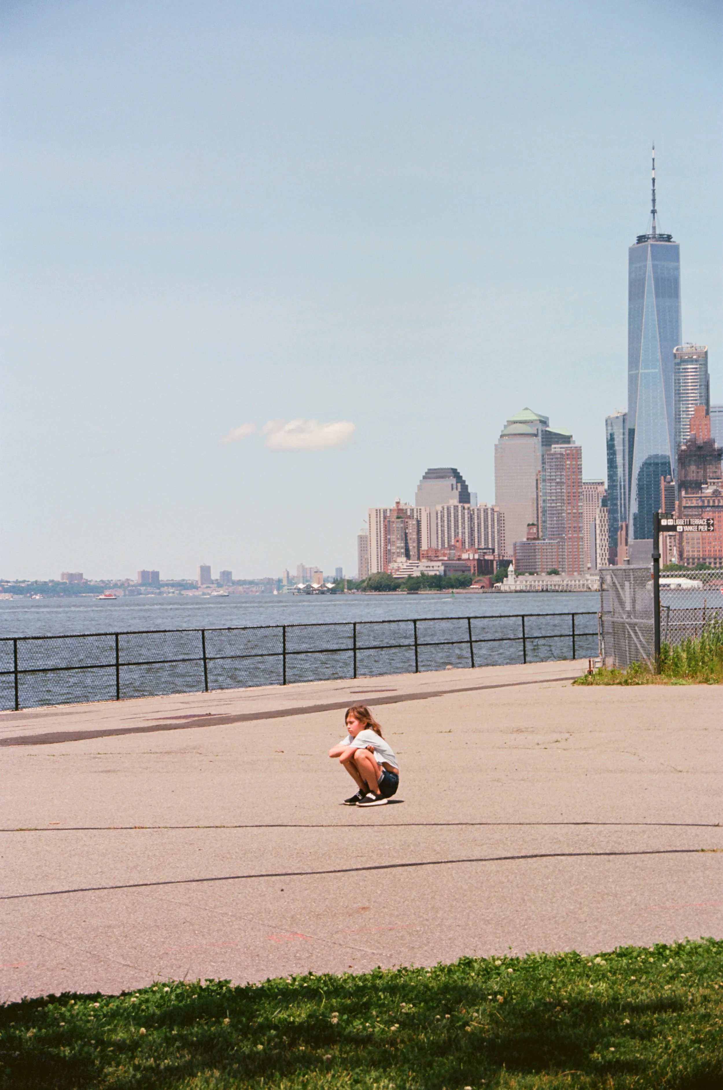 A girl squatting on a paved outdoor area beside a green hedge, with a city skyline including the One World Trade Center in the background. scenic temper tantrum. 