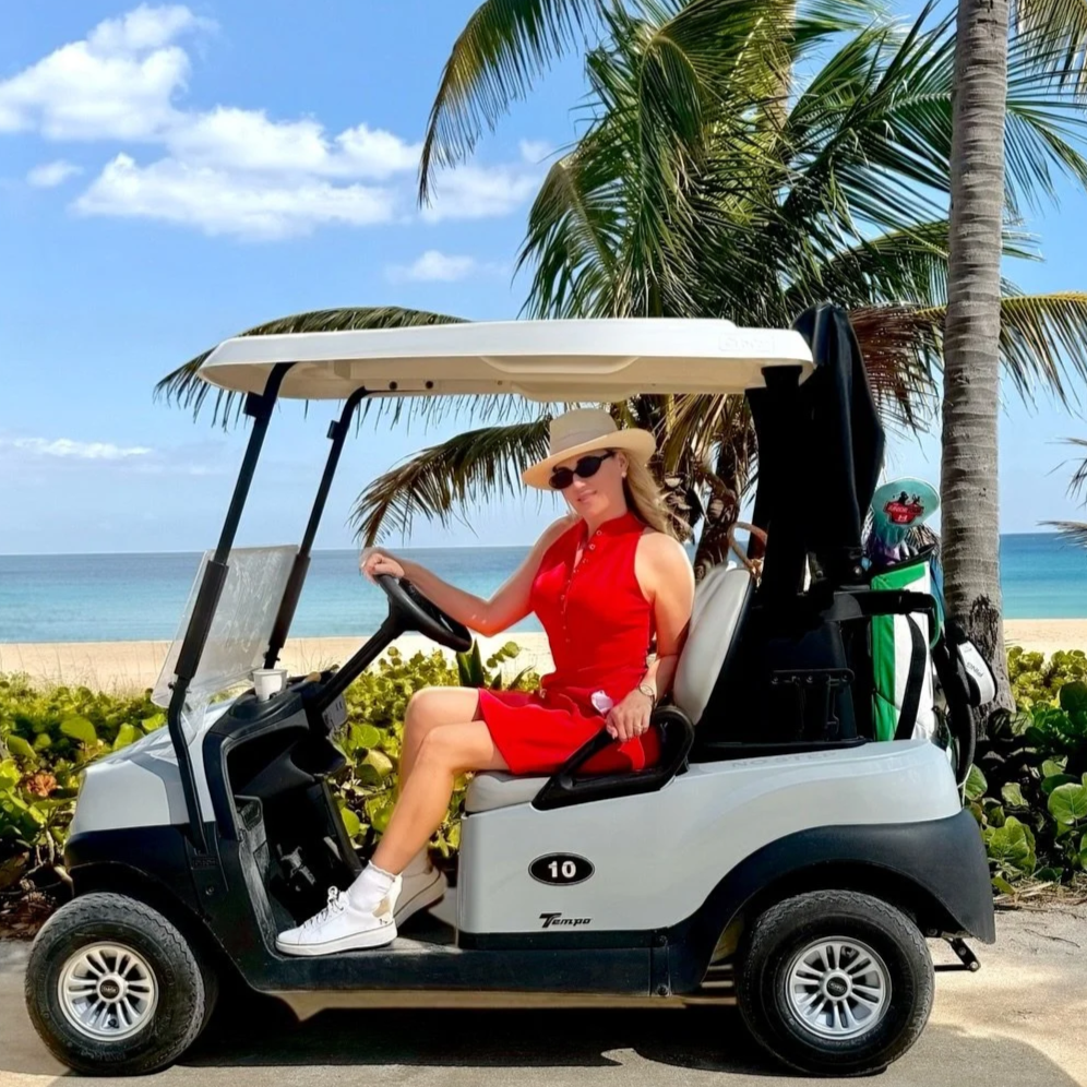 Woman in red dress and hat sitting in a golf cart on a beach with palm trees and ocean in the background.