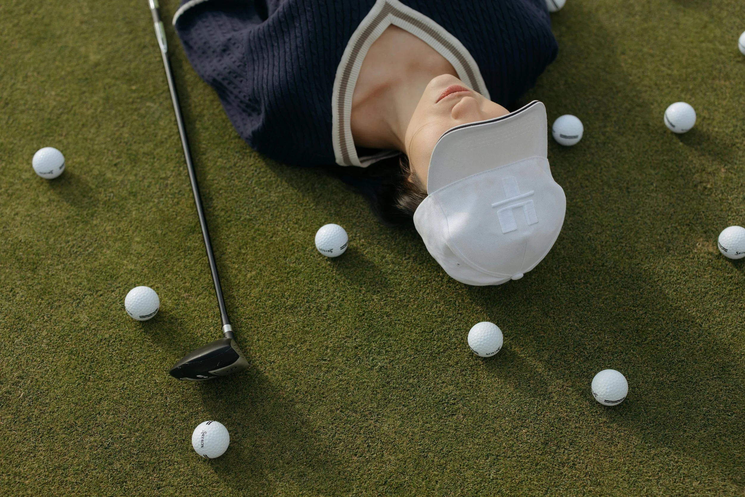 A woman lying on a golf course with a golf club beside her, surrounded by golf balls.