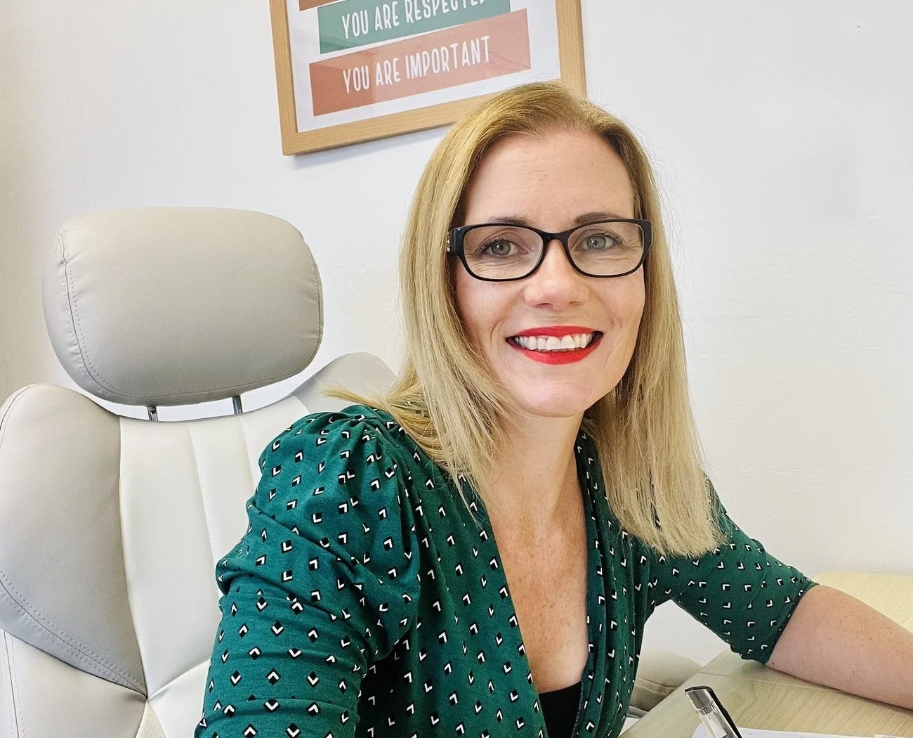 A woman with blonde hair, wearing black glasses and red lipstick, smiling while sitting in a white office chair with an adjustable headrest, in an office with a white wall and a colorful framed quote in the background.