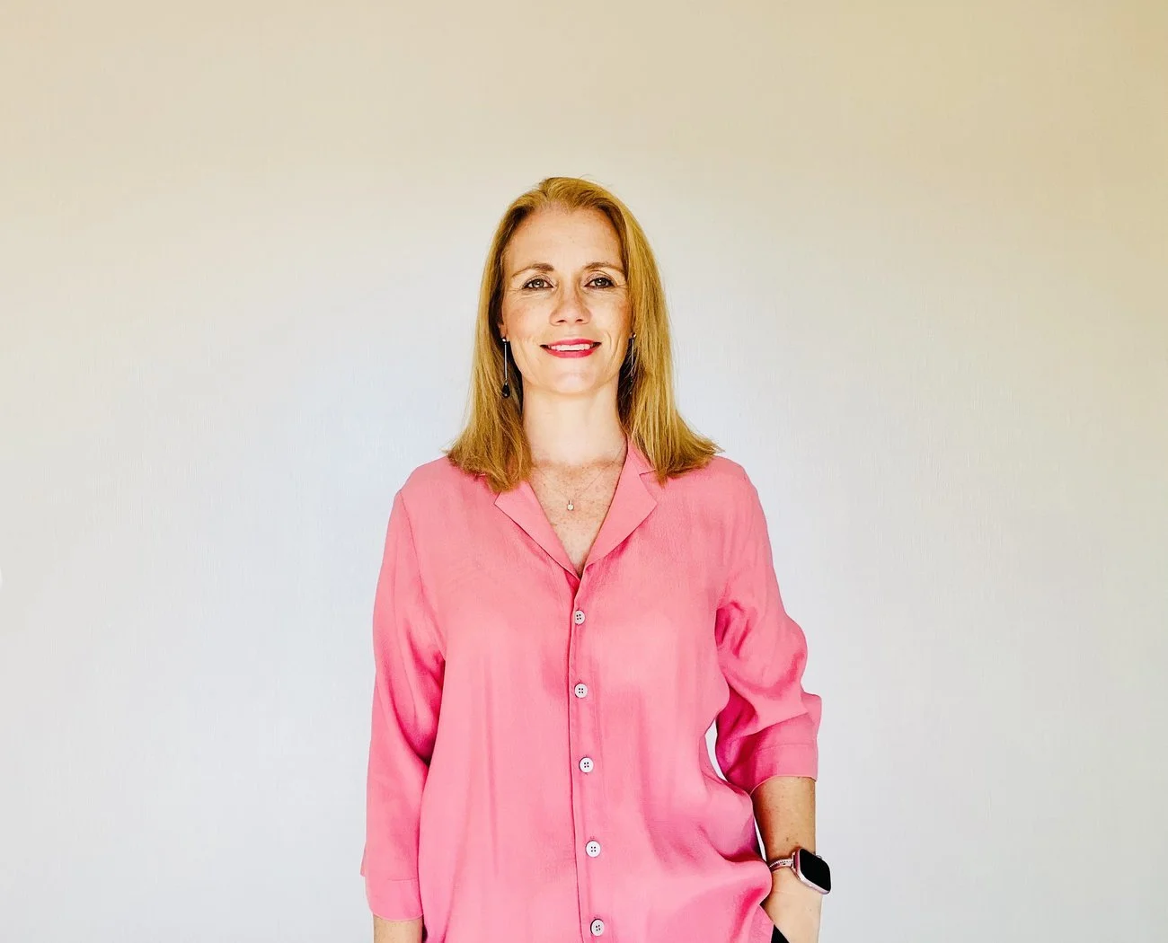 A woman with shoulder-length blonde hair, wearing a pink button-up shirt, standing against a plain light-colored wall. She is smiling and wearing a smartwatch and earrings.