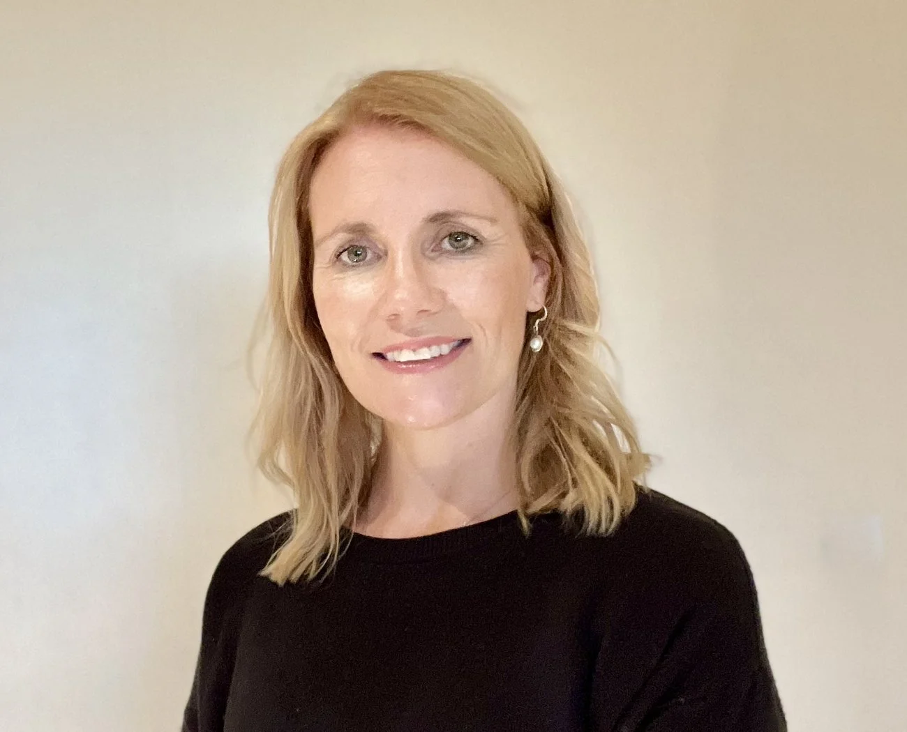 A woman with shoulder-length blonde hair, smiling, wearing a black top and pearl earrings, standing against a light-colored wall.