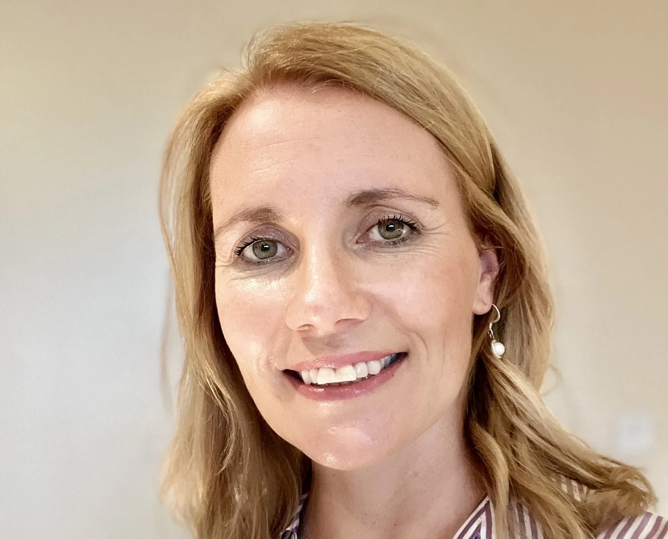 Close-up of a smiling woman with shoulder-length blonde hair, green eyes, and wearing a striped shirt and pearl earring.