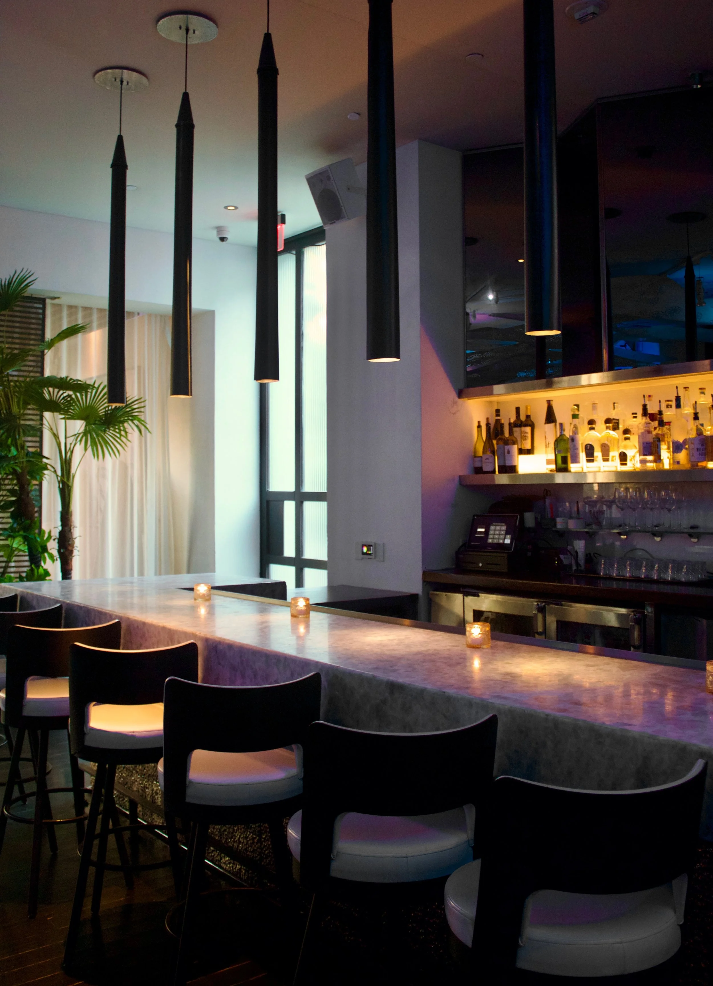 A modern bar interior with hanging black pendant lights, a long marble counter with small candles, a row of white chairs, a bar with various bottles, and a plant near a tall window.