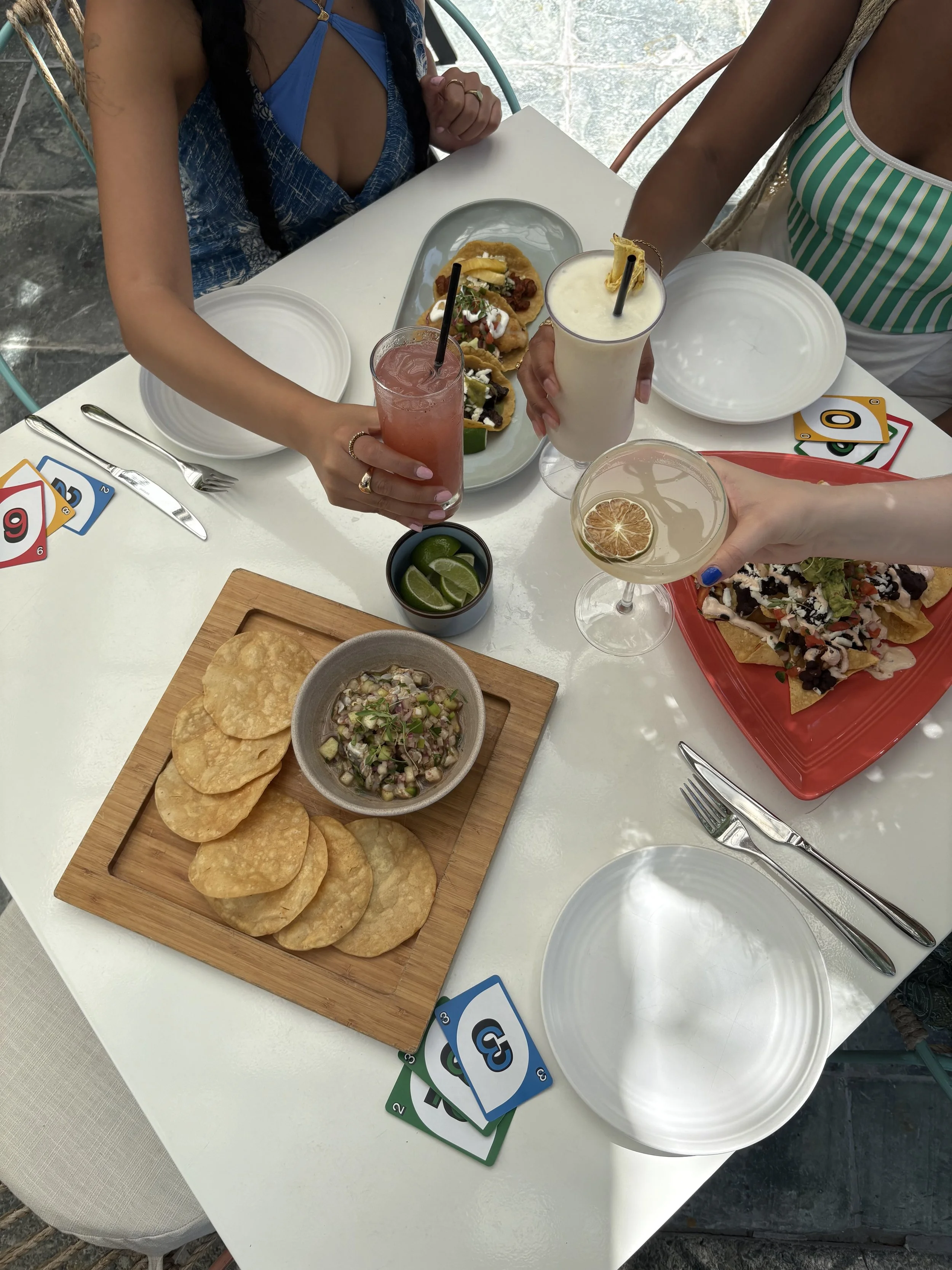 Table with Mexican food and drinks, including tacos on a platter, a bowl of lime wedges, and tortilla chips with salsa, along with playing cards.