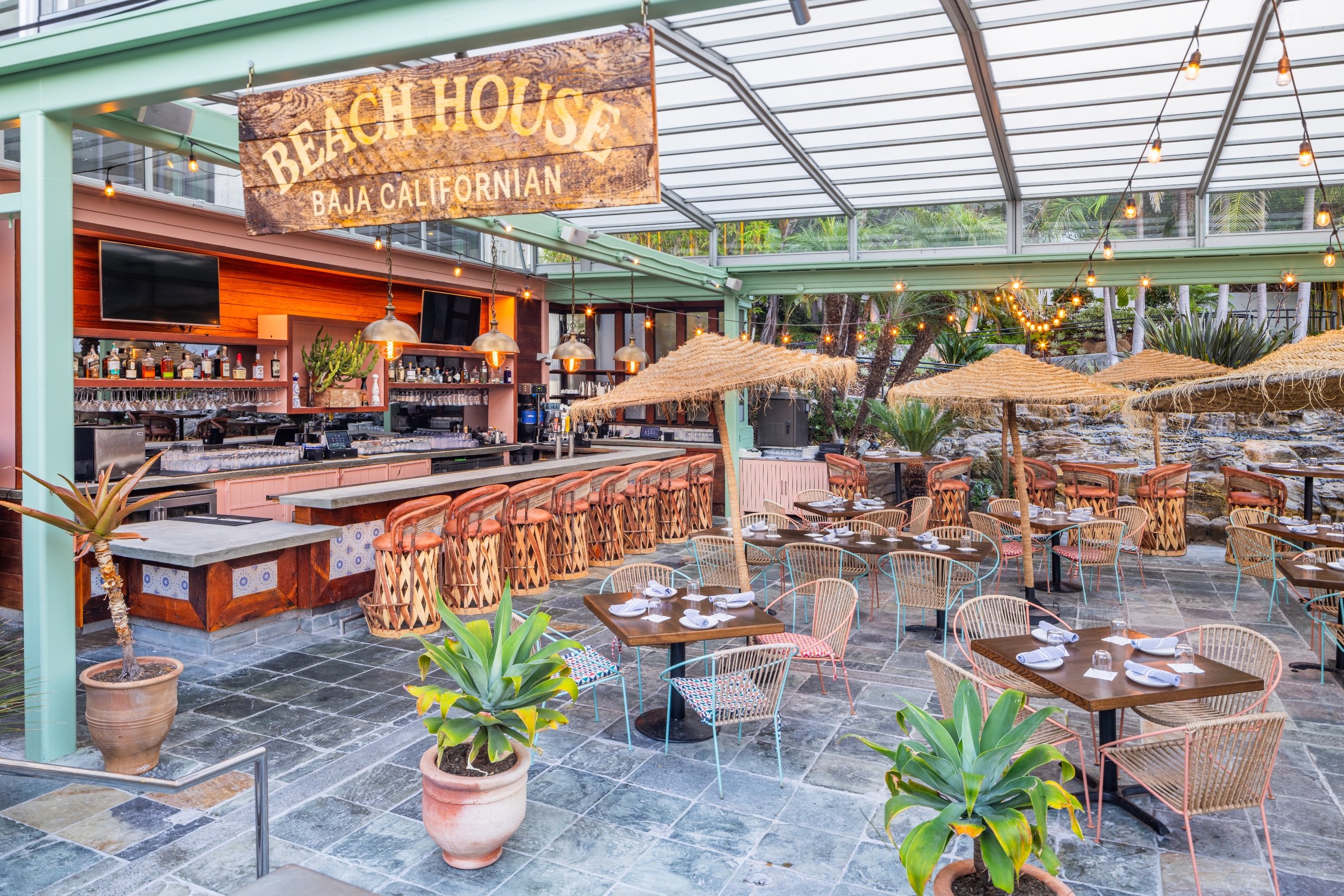 Outdoor restaurant with wooden tables, chairs, potted plants, string lights, a bar area with bottles, and a bamboo shaded patio with a sign reading 'Beach House Baja Californian'.
