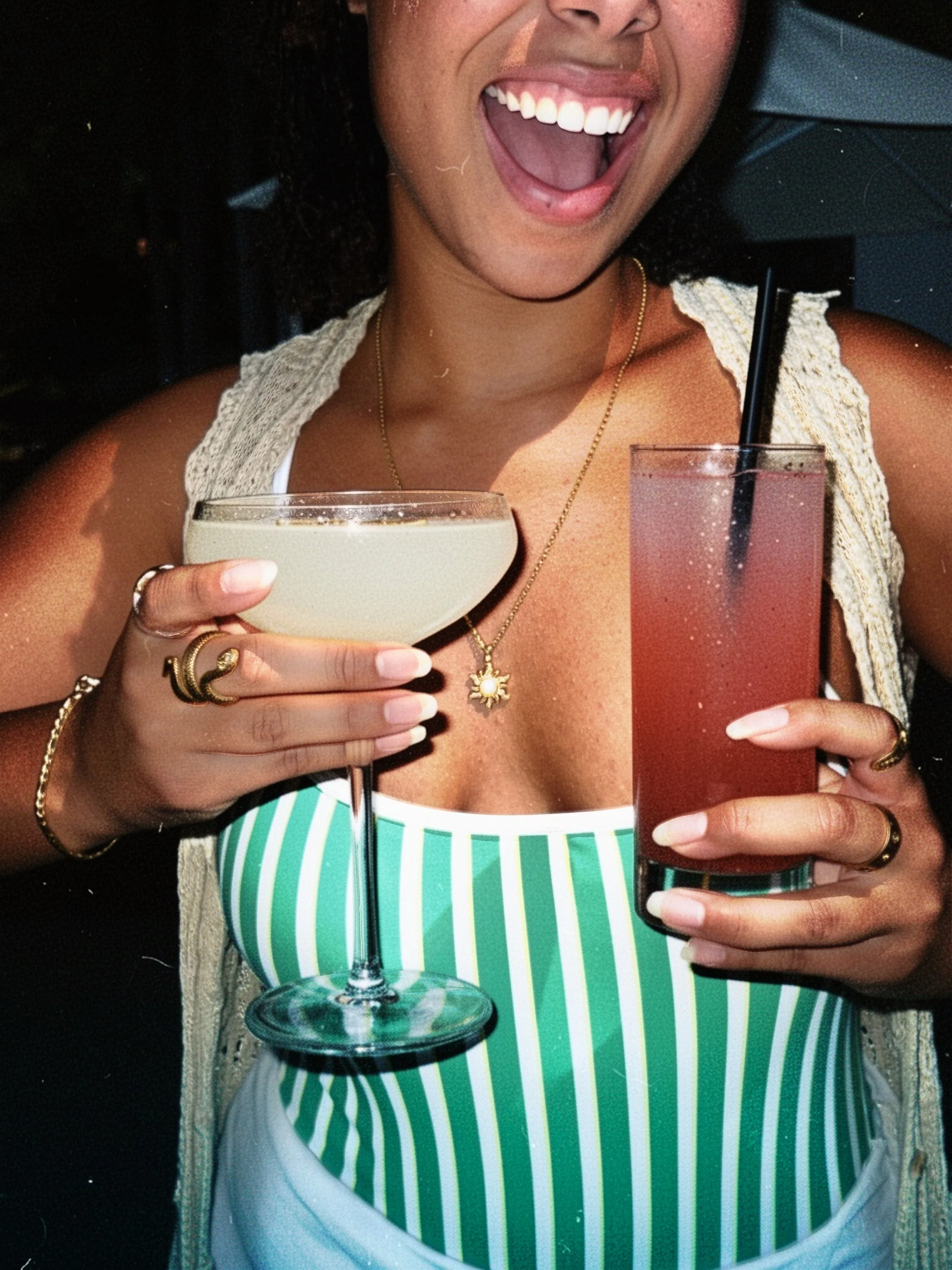 A woman smiling with an open mouth, holding two colorful drinks, one in each hand. She is wearing a green and white striped top, layered necklaces, and rings.