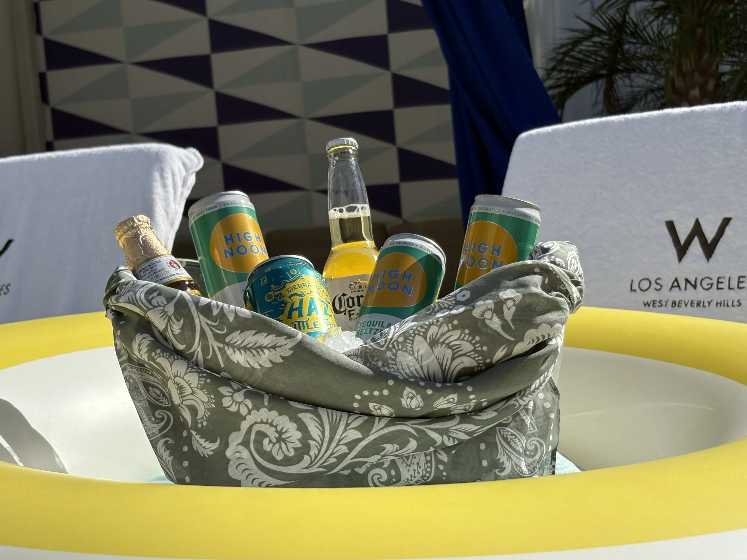 A pedestal basket of assorted canned beverages and bottled drinks on ice, inside a boat with yellow and white interior, with a geometric patterned wall and some greenery in the background.