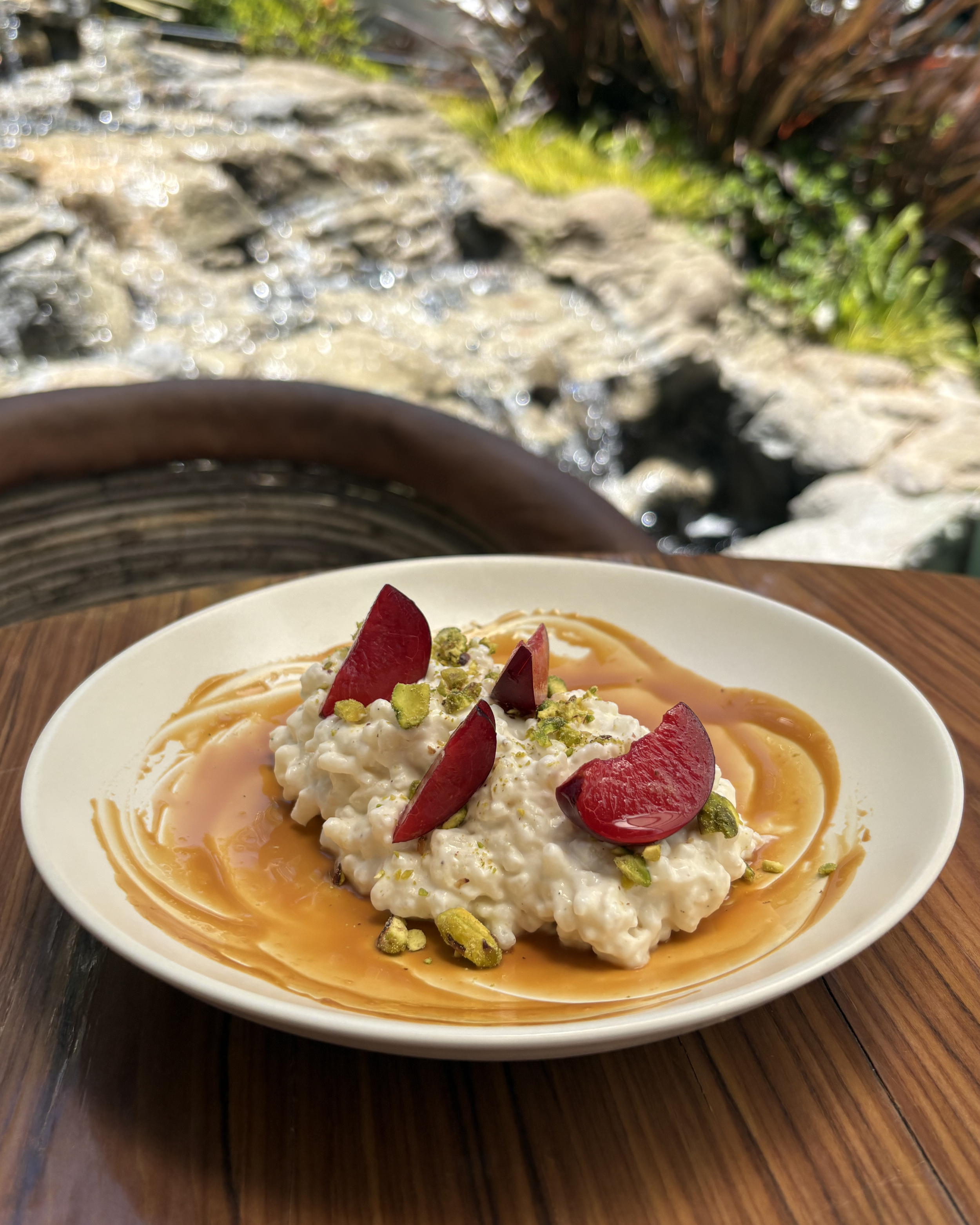 A bowl of ricotta cheese garnished with sliced strawberries and chopped pistachios, drizzled with honey, on a wooden table outdoors.