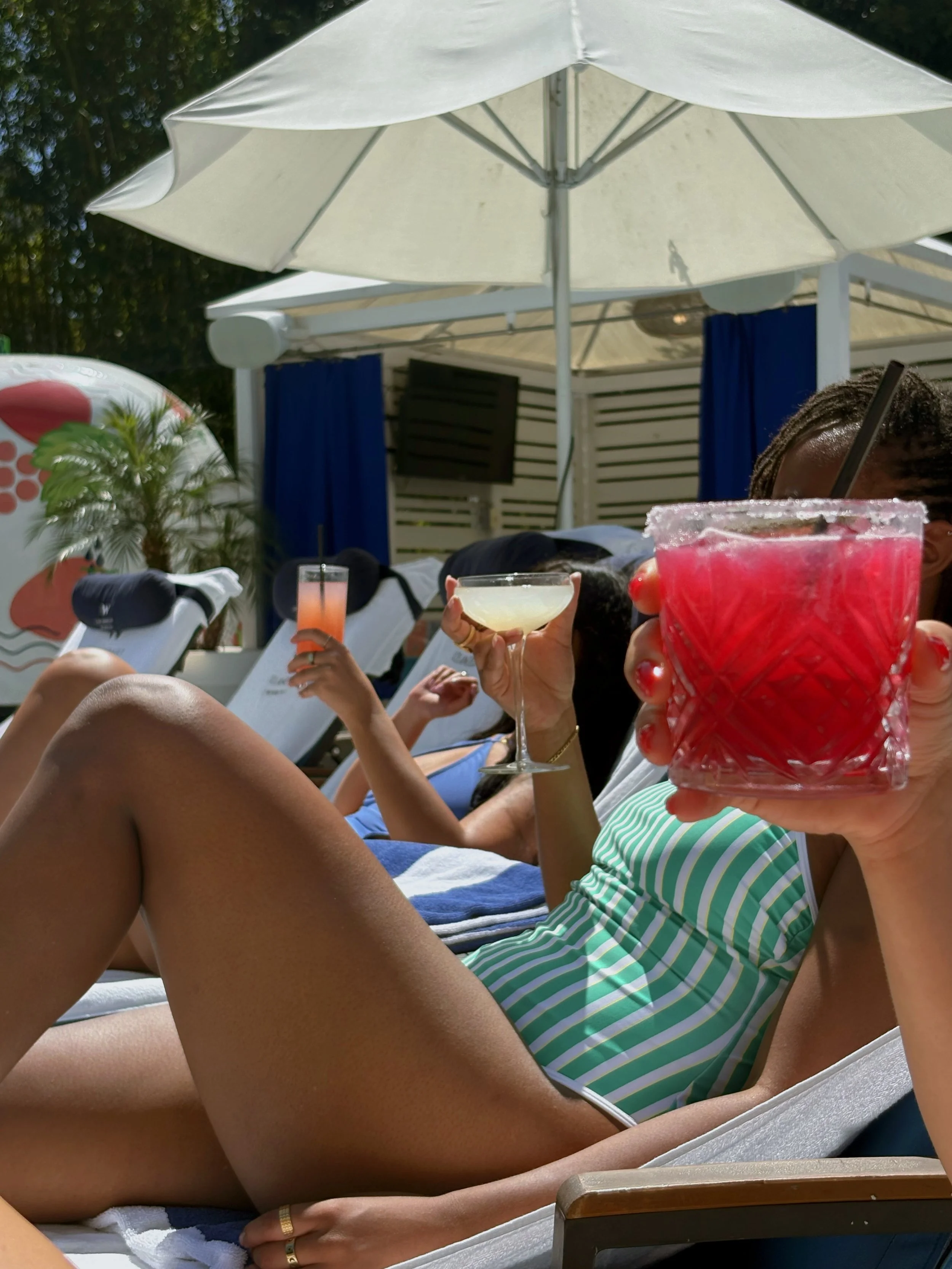 People relaxing poolside on lounge chairs under umbrellas, holding drinks, with a woman in green and white striped swimwear holding a red cocktail.
