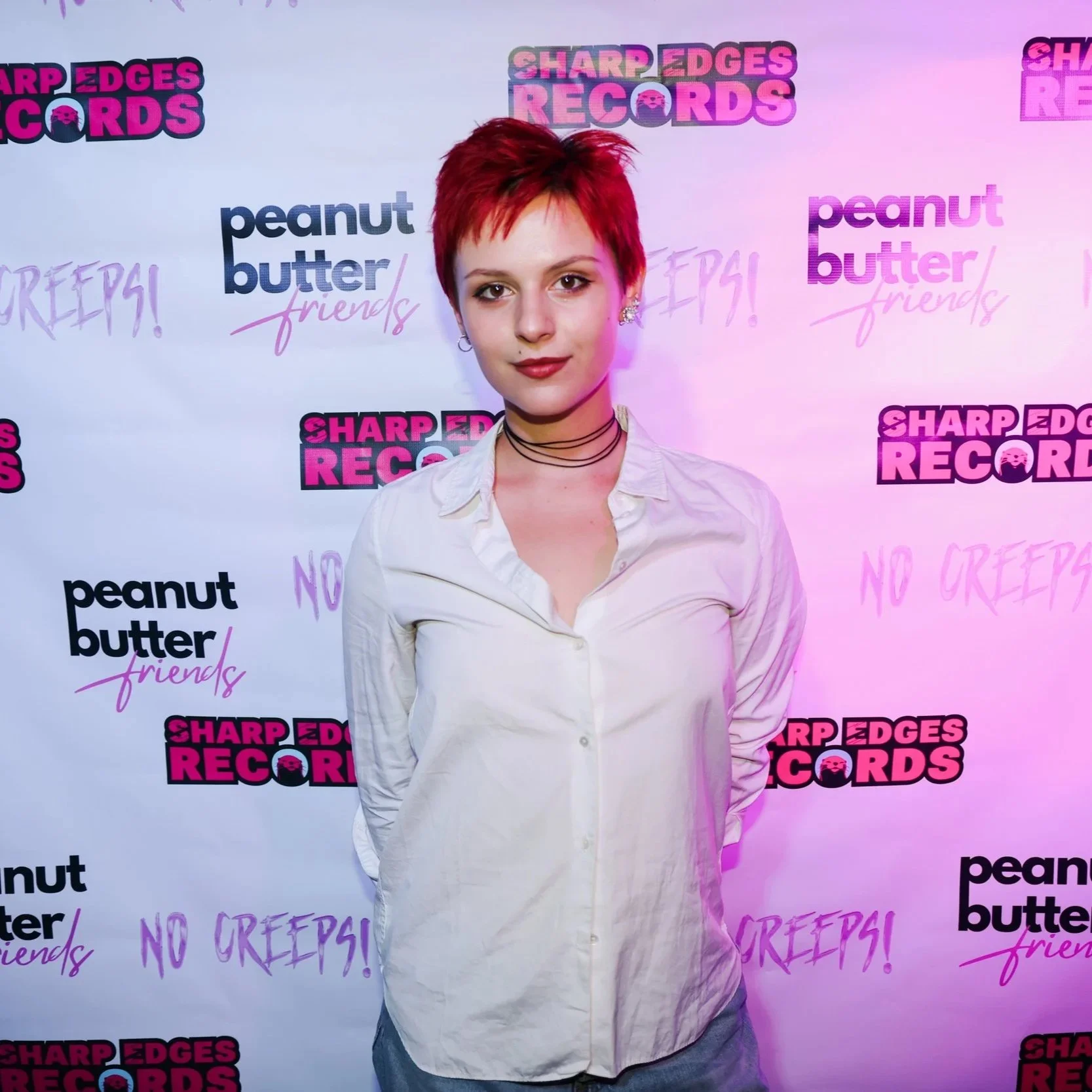 Young woman with short red hair and earrings, wearing a white shirt, standing in front of a backdrop with text and logos for Sharp Edges Records, Peanut Butter Friends, and No Gripe.