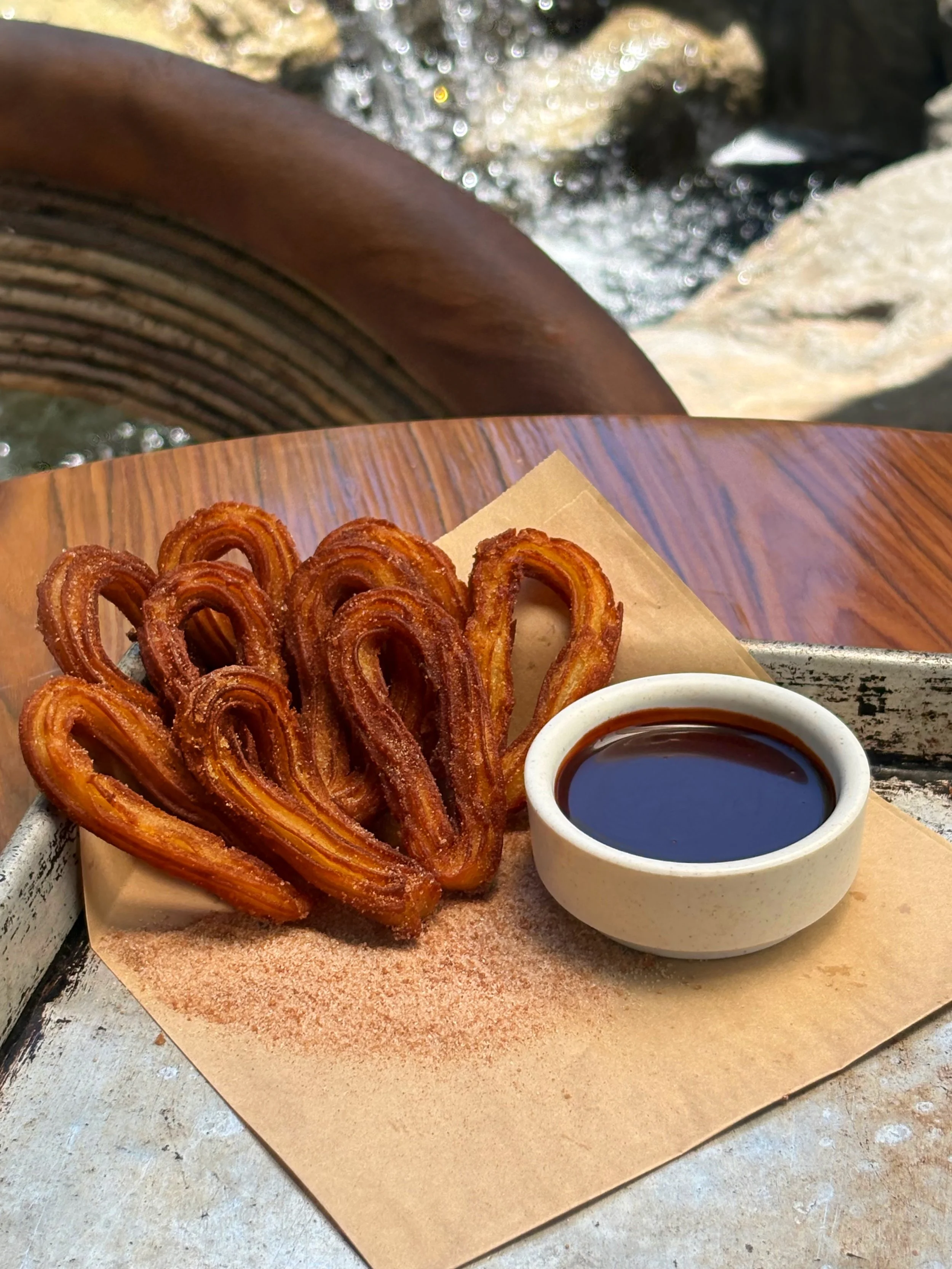 Crispy churros with a side of chocolate sauce on a wooden tray with a printed paper liner, placed on a rustic metal surface near flowing water.