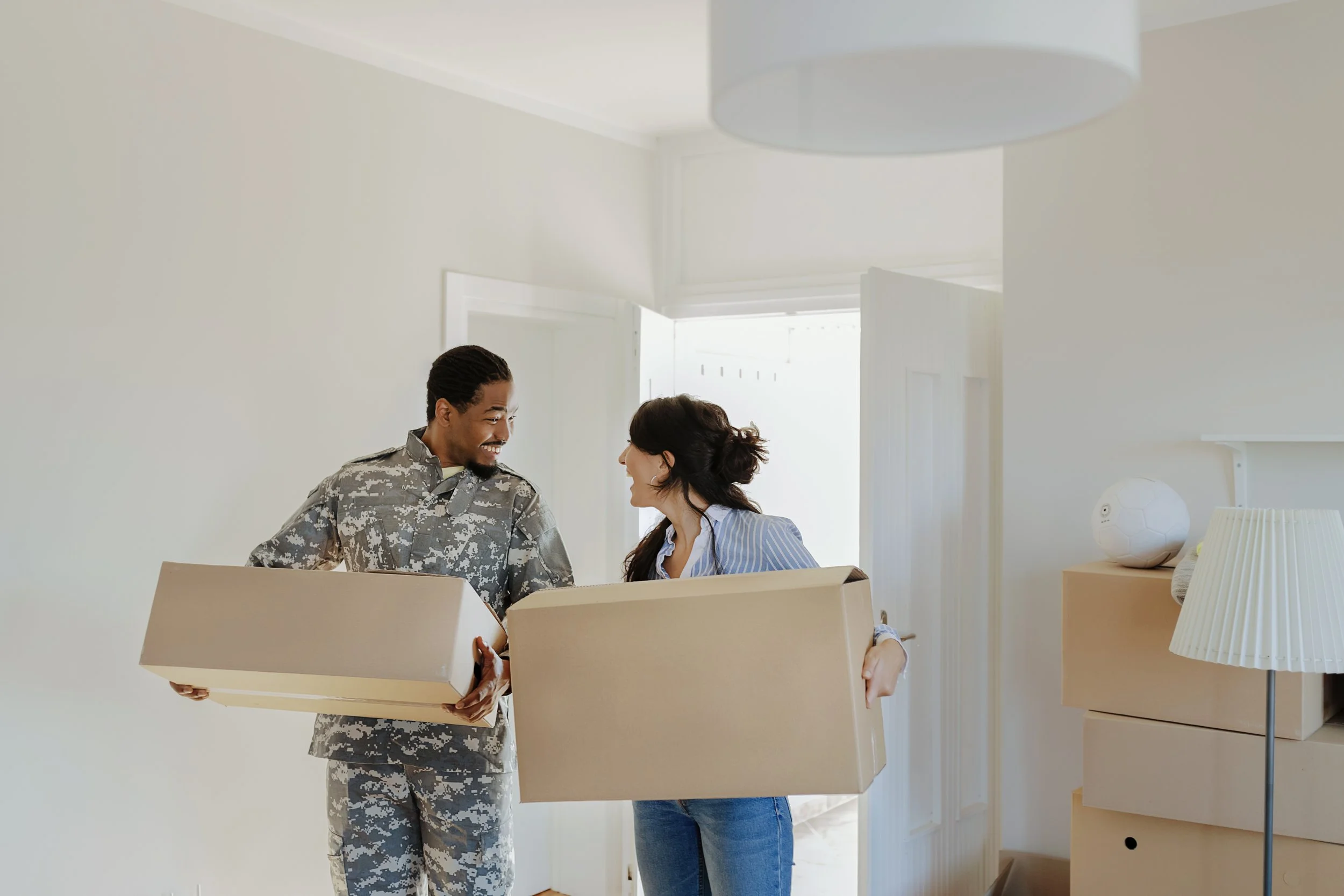 A soldier and a woman are smiling and looking at each other while holding an open cardboard box in a well-lit room. The room has a closed door, a standing lamp, and a decorative object resembling a soccer ball on a piece of furniture.
