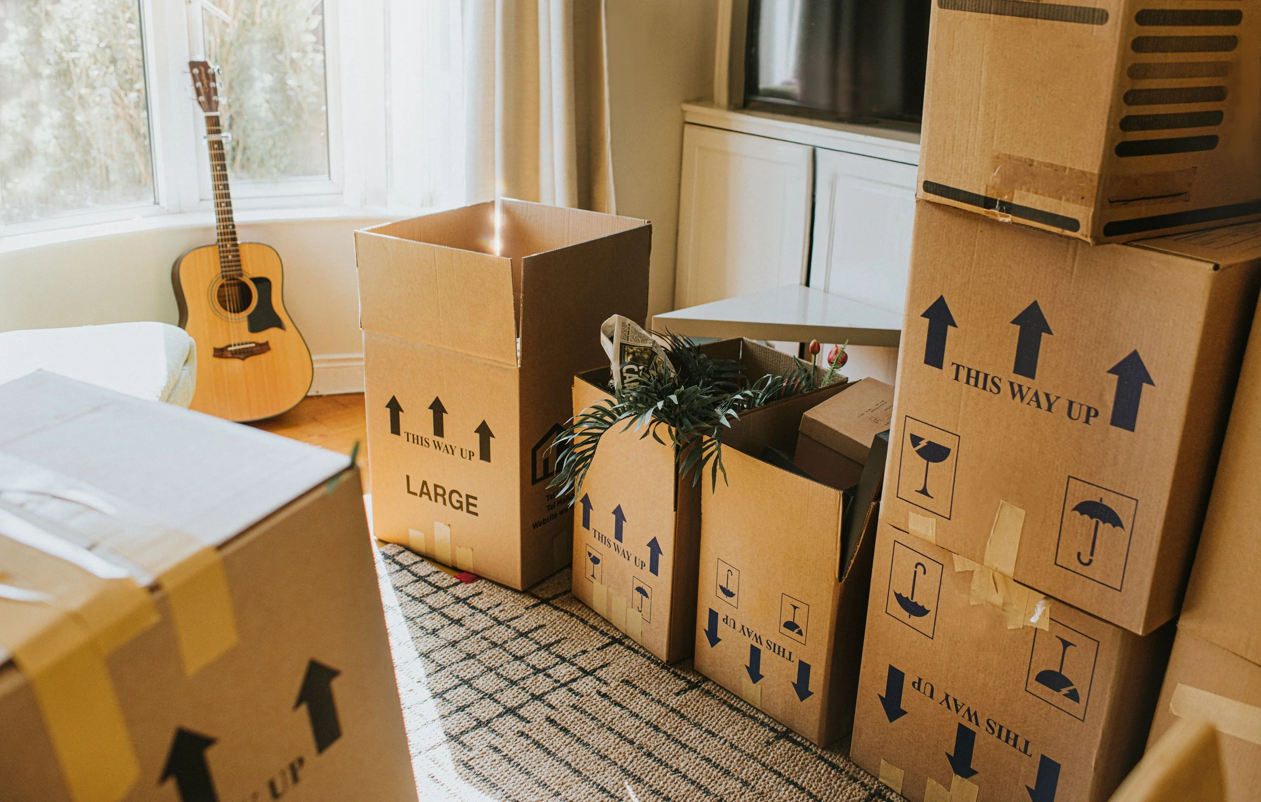 Room with moving boxes, some marked 'This Way Up,' a guitar leaning against a window, and a TV on a cabinet.