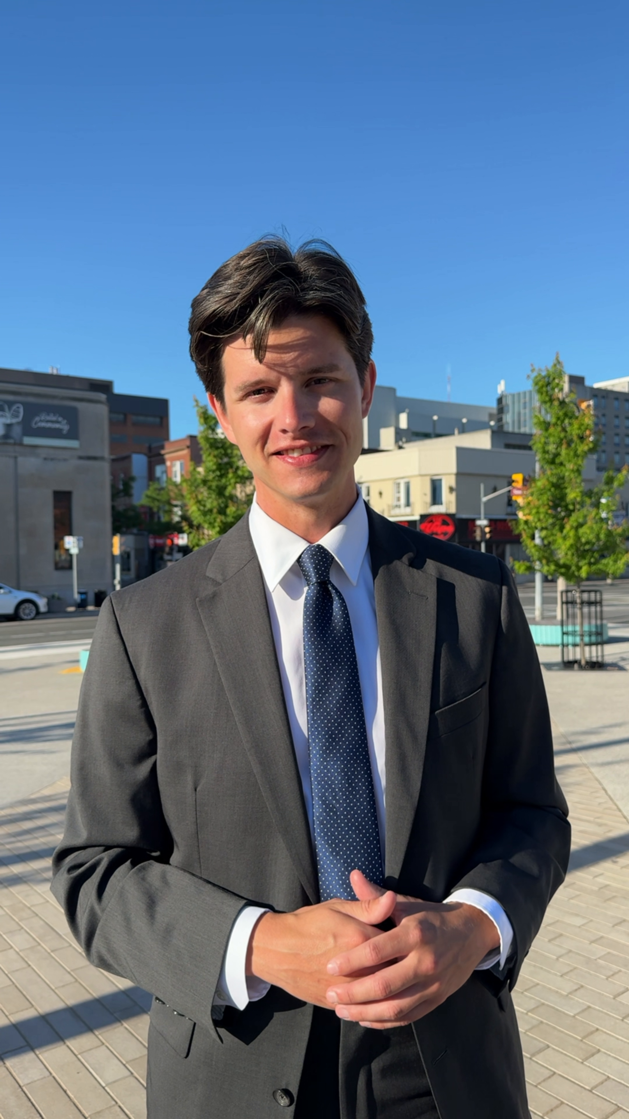 Young man in a gray suit and blue polka dot tie standing outdoors on a city sidewalk, with office buildings, trees, and a clear blue sky in the background.