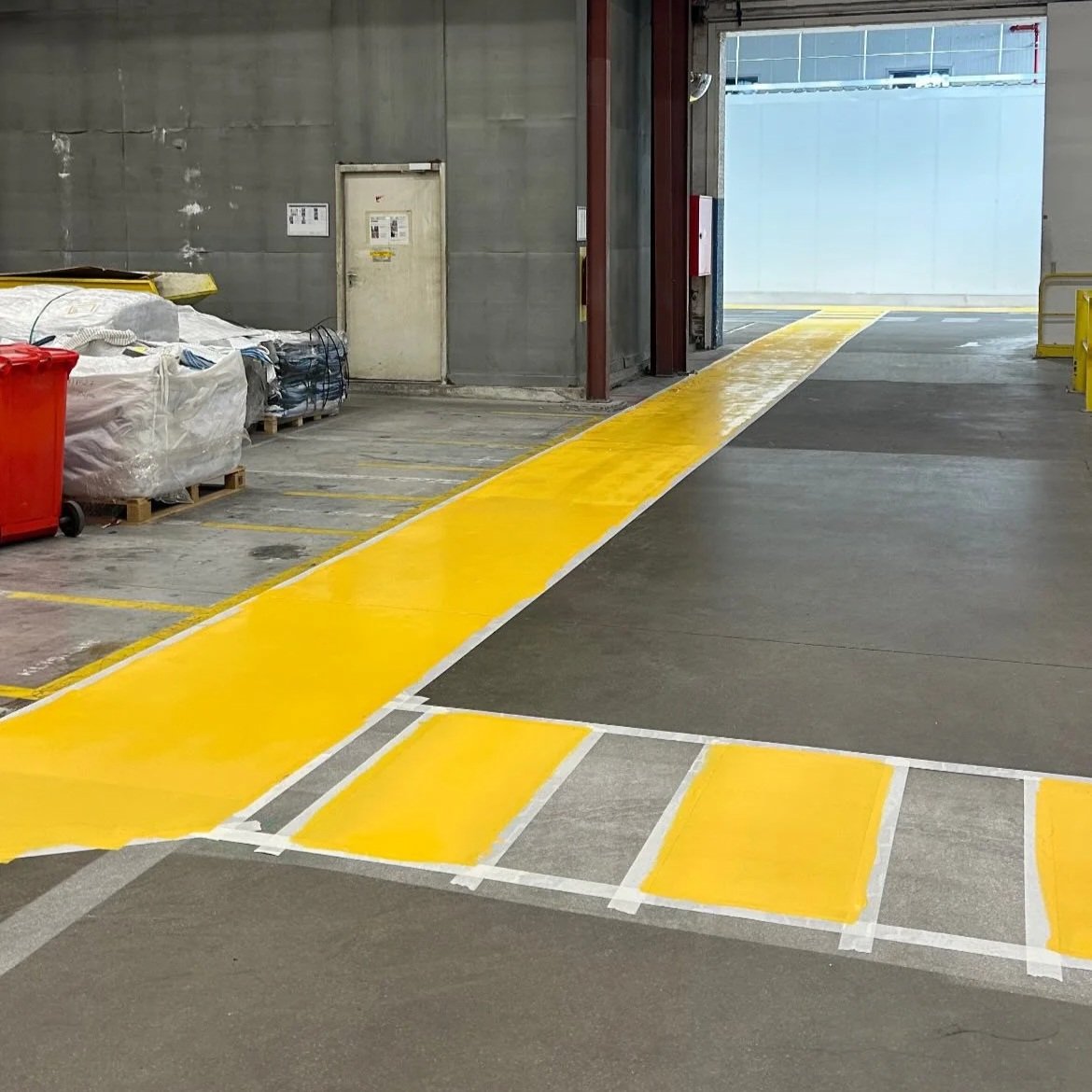 Gele verfmarkering en looproute in een parkeerplaats of magazijn, met pallet met plastic verpakkingen en een rode afvalcontainer links.